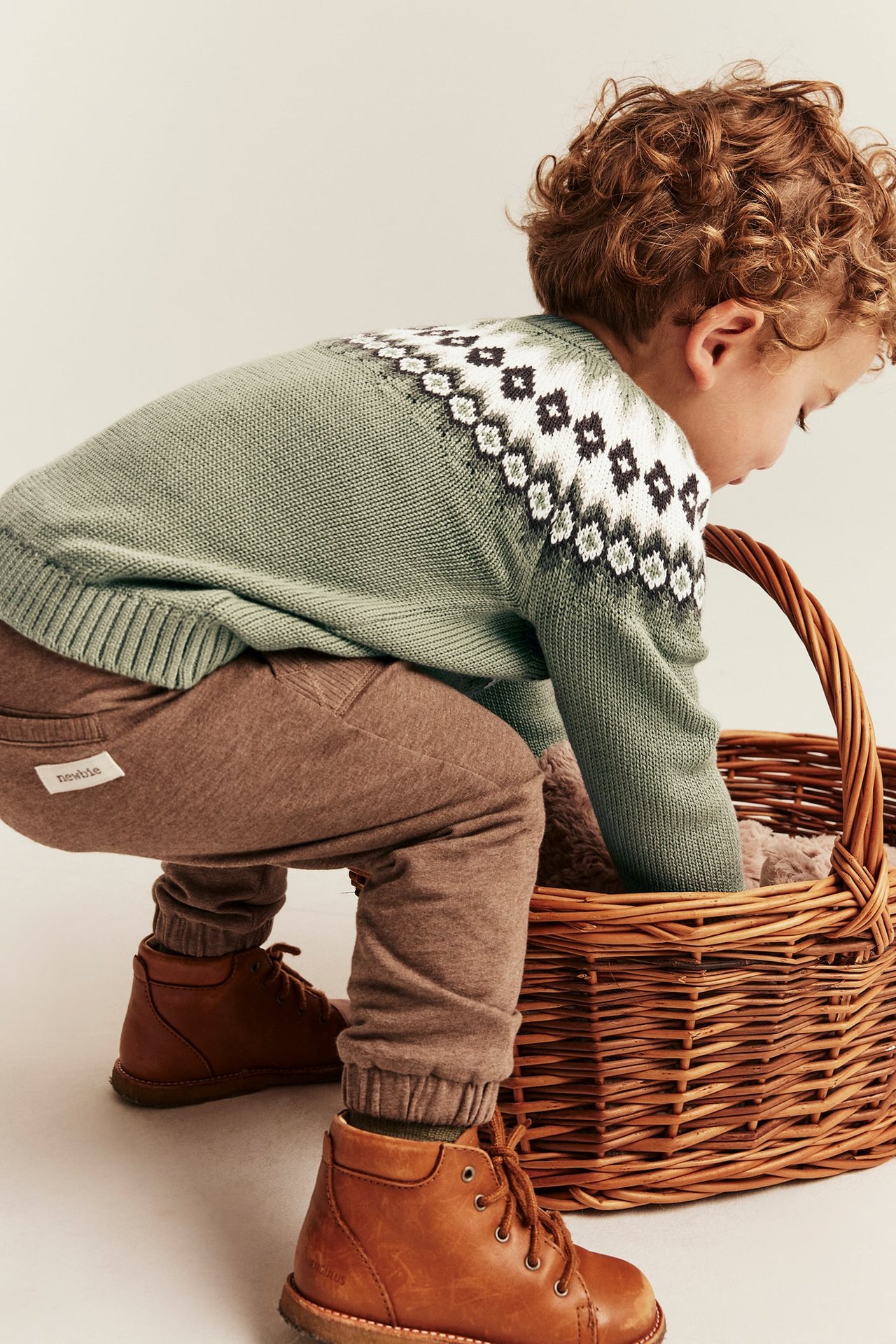 Back view of kid in green patterned knit sweater, brown joggers, brown leather boots, reaching into a basket.