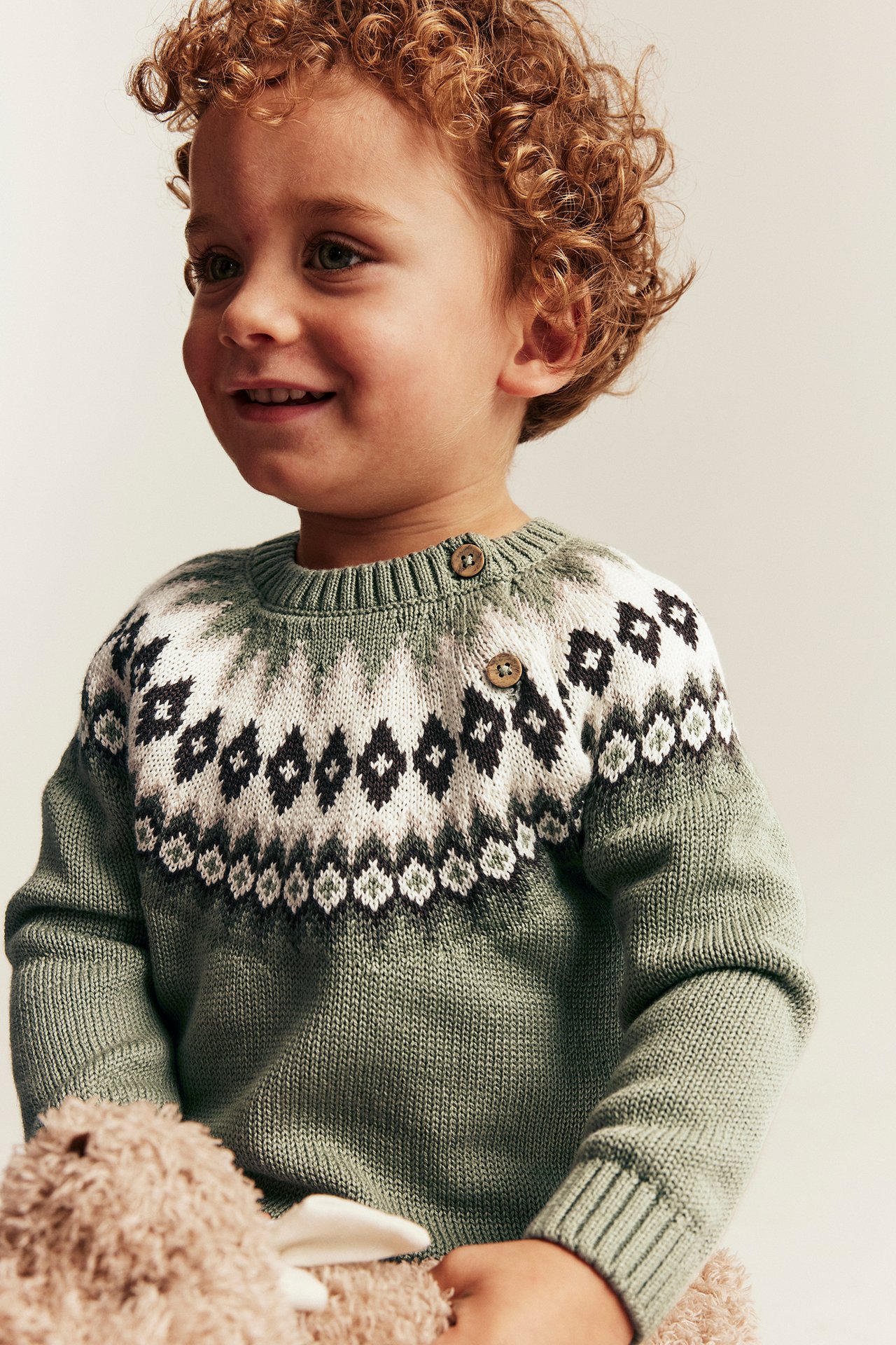 Front view of a kid in a sage green Fair Isle knitted sweater with buttons, holding a plush toy.
