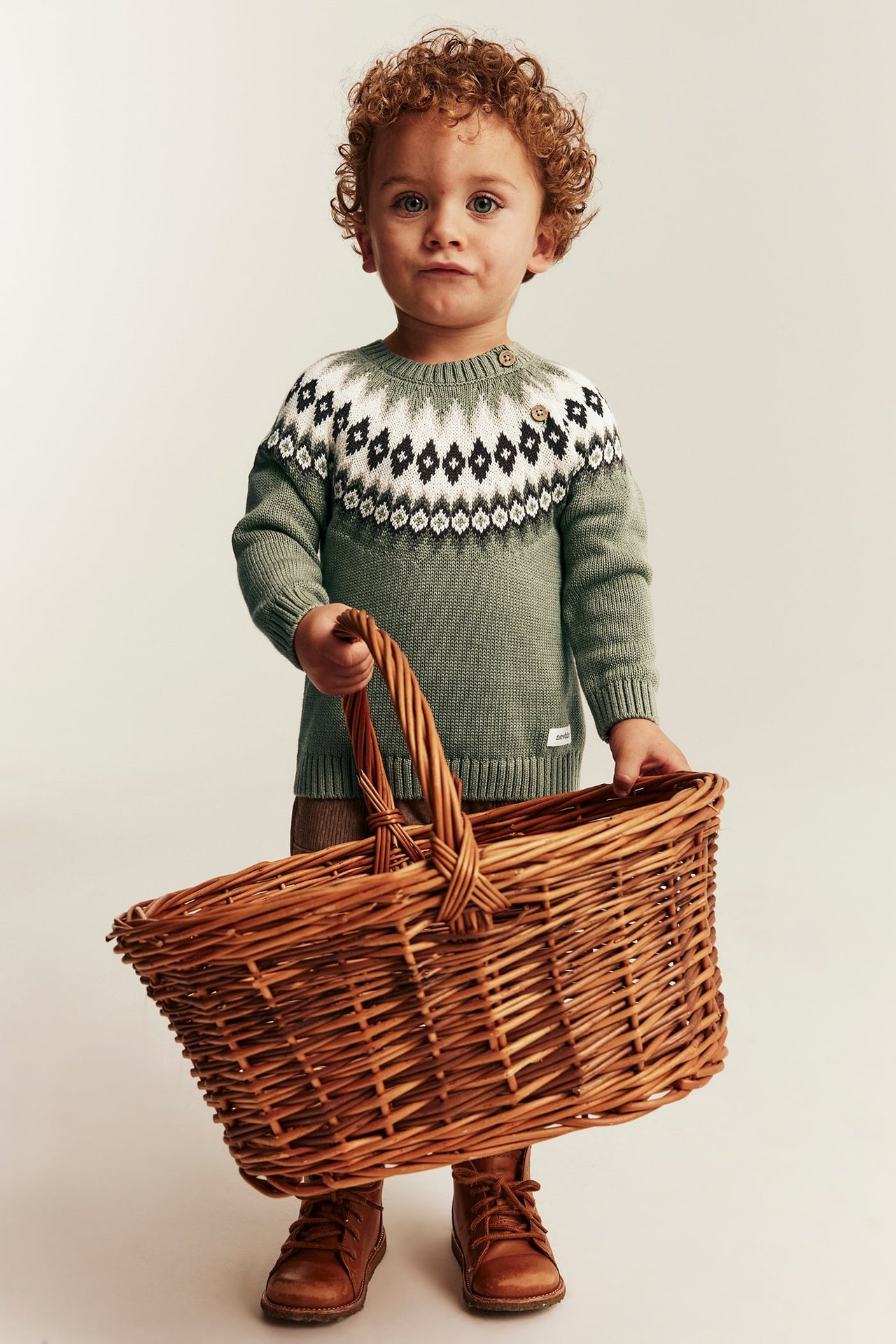 Kid in green fair isle sweater with black and white, brown pants, boots, holding wicker basket.