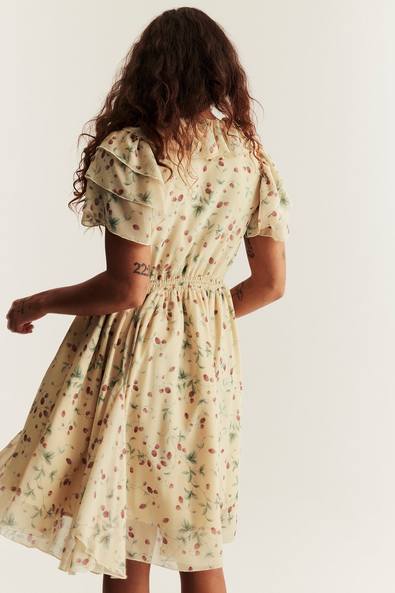 Back view of a woman wearing a yellow patterned chiffon dress with ruffled sleeves and an elasticated waist.