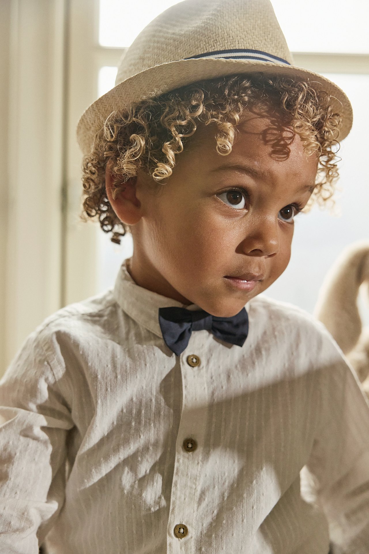 Close-up of kid in light beige straw hat with blue striped band, light shirt, and navy bow tie.