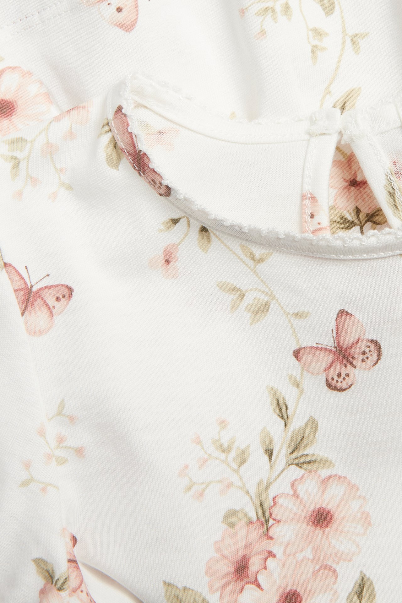 Nahaufnahme: Offwhite-Baumwoll-Jersey-Top mit rosa Blumen- und Schmetterlingsmuster, Picot-Ausschnitt und Knopfverschluss hinten.