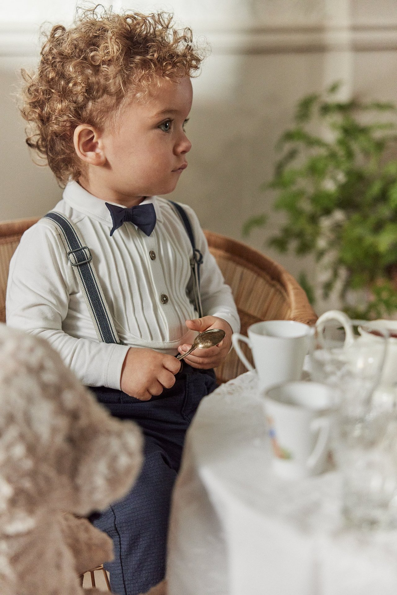 Side view of a baby wearing an off-white pleated bodysuit, dark blue bow tie, trousers, and suspenders.