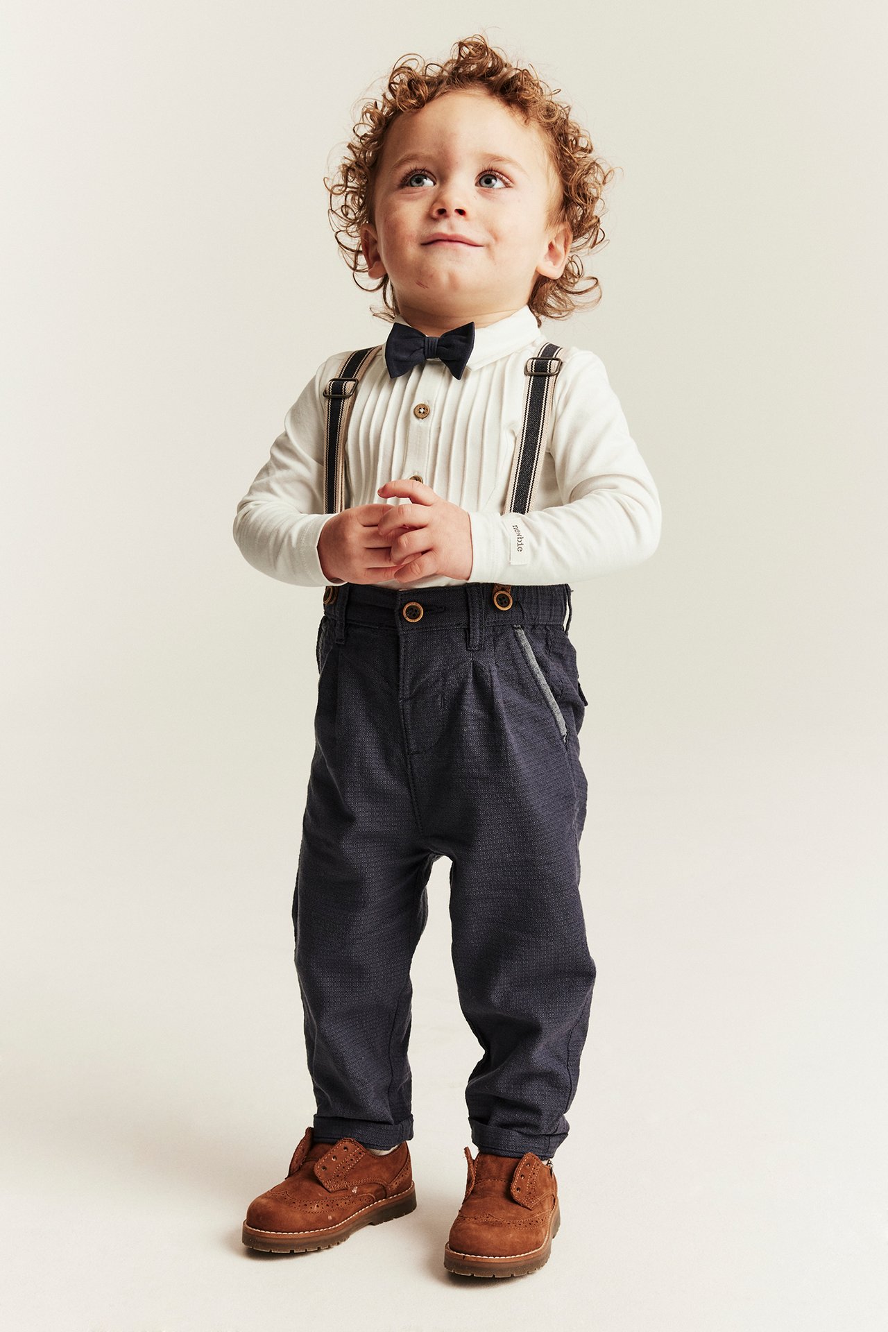 Front view: Baby boy in off-white pleated bodysuit, dark bow tie, blue trousers, suspenders, brown shoes.