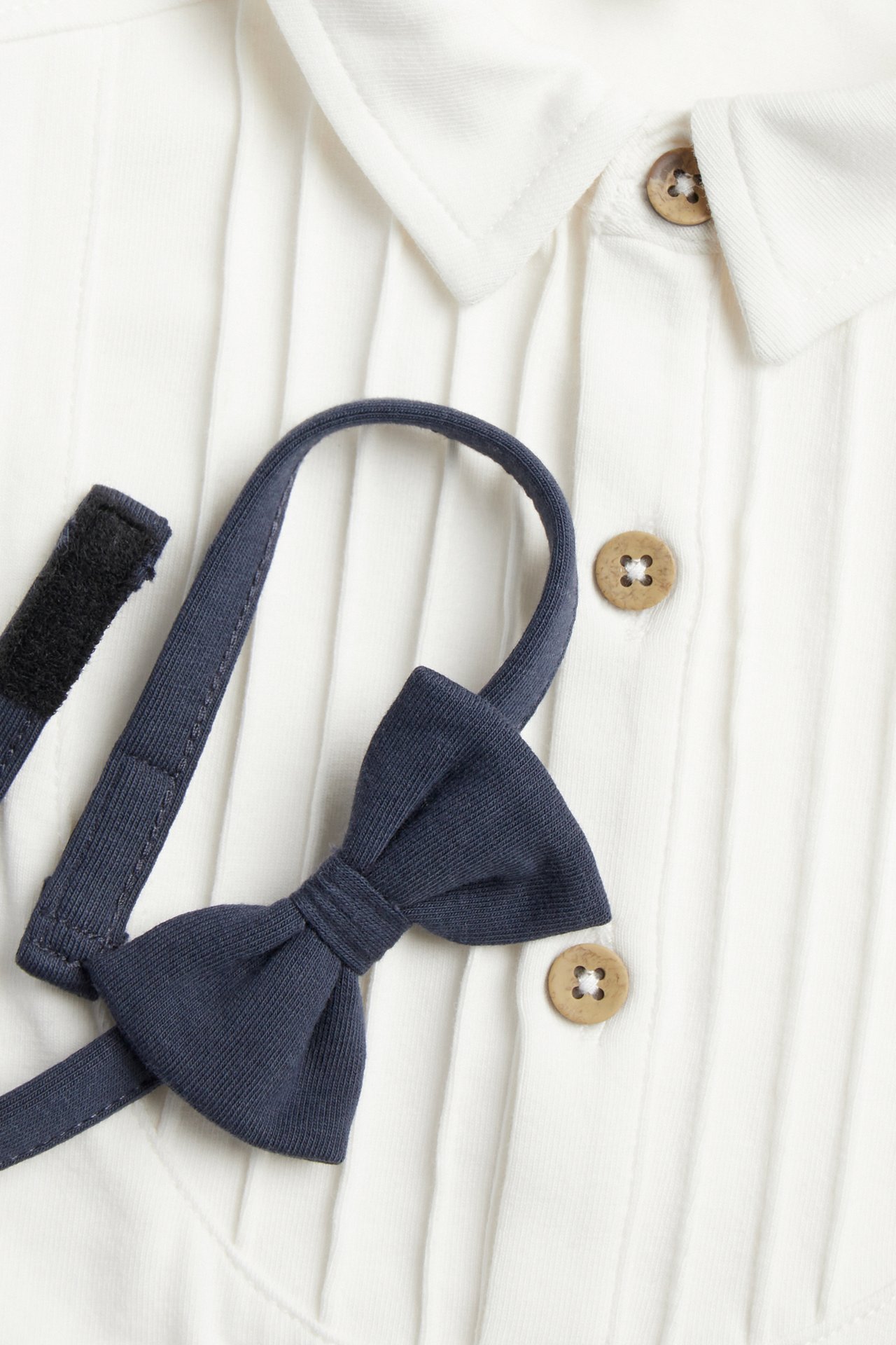 Close-up front view of an off-white long-sleeved bodysuit with pintucks, wooden buttons, and a dark blue detachable bow tie.