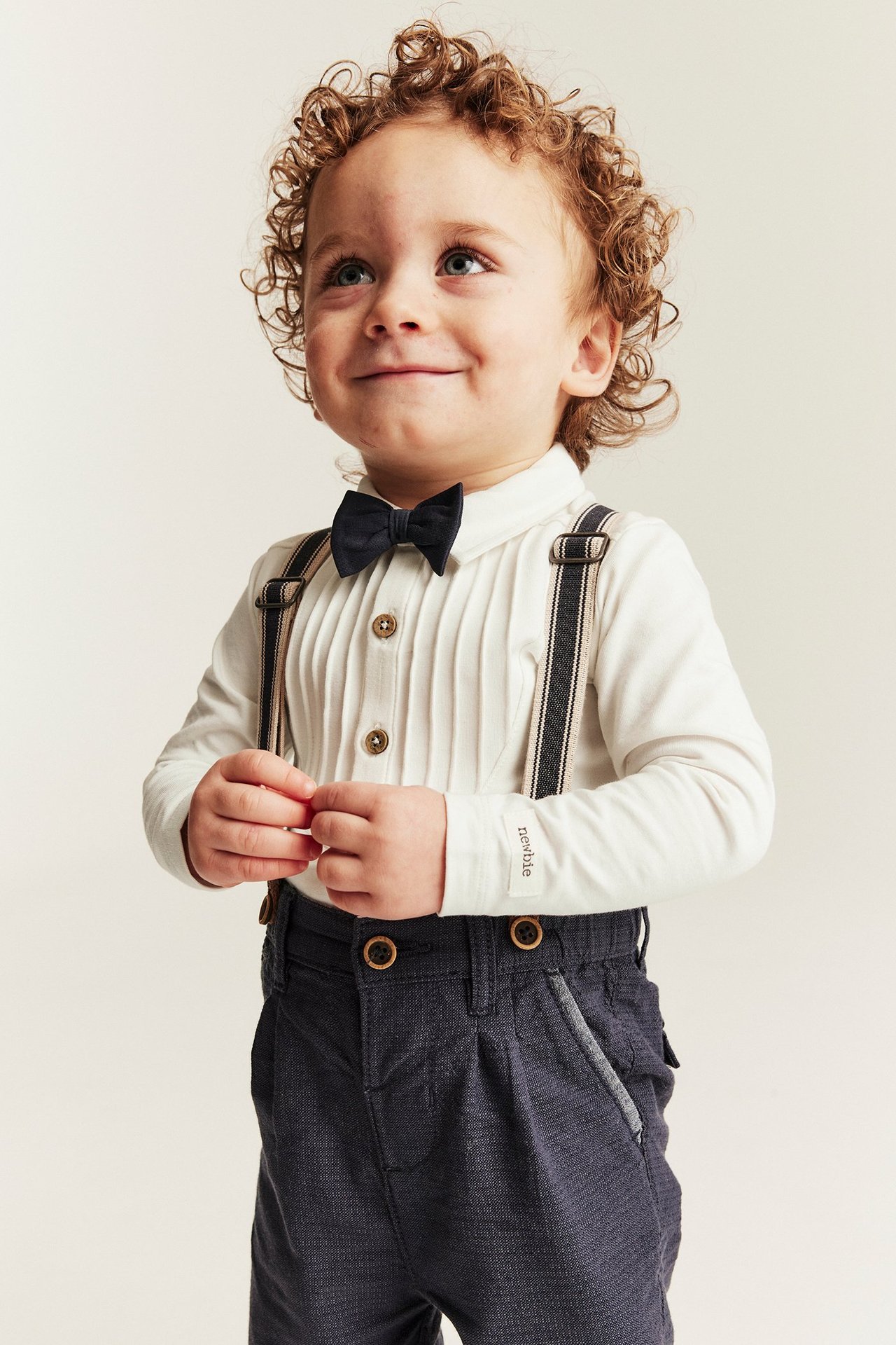 Front view: Baby in off-white pintucked bodysuit, dark blue bow tie, striped suspenders and dark grey trousers.
