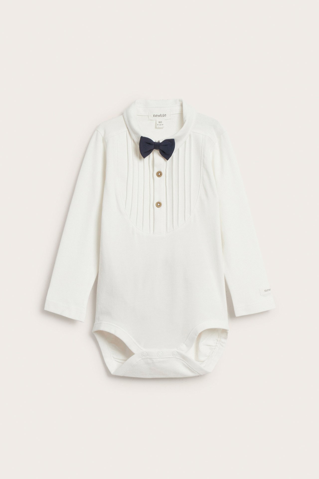 Front view of an off-white long-sleeved baby bodysuit with a collar, pintucks, and a dark blue detachable bow tie.
