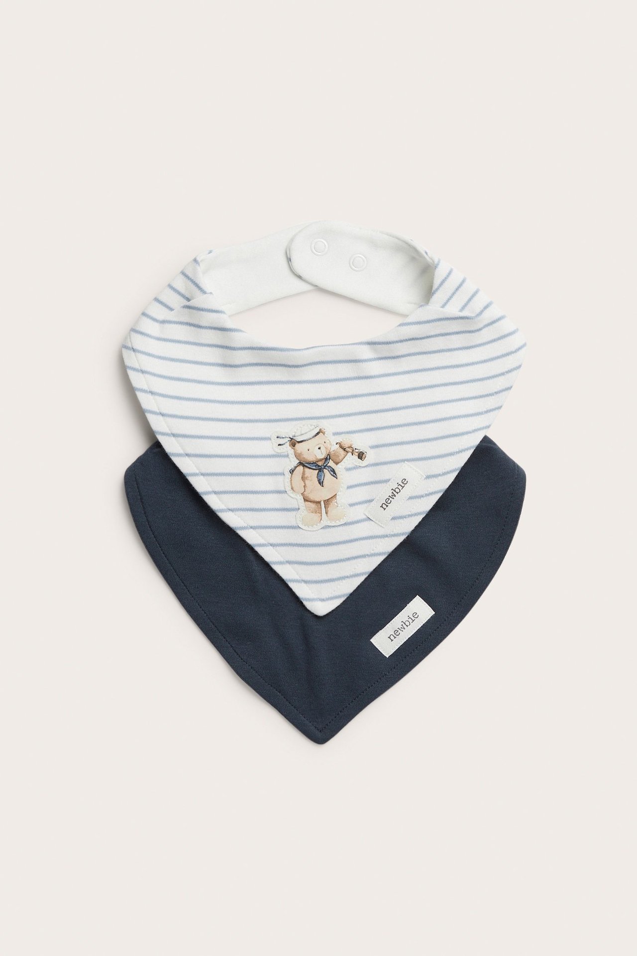 Vorderansicht: Zwei Baumwoll-Bandana-Lätzchen. Eines gestreift cremeweiß mit Seebären-Applikation, eines uni marineblau, mit Druckknöpfen.