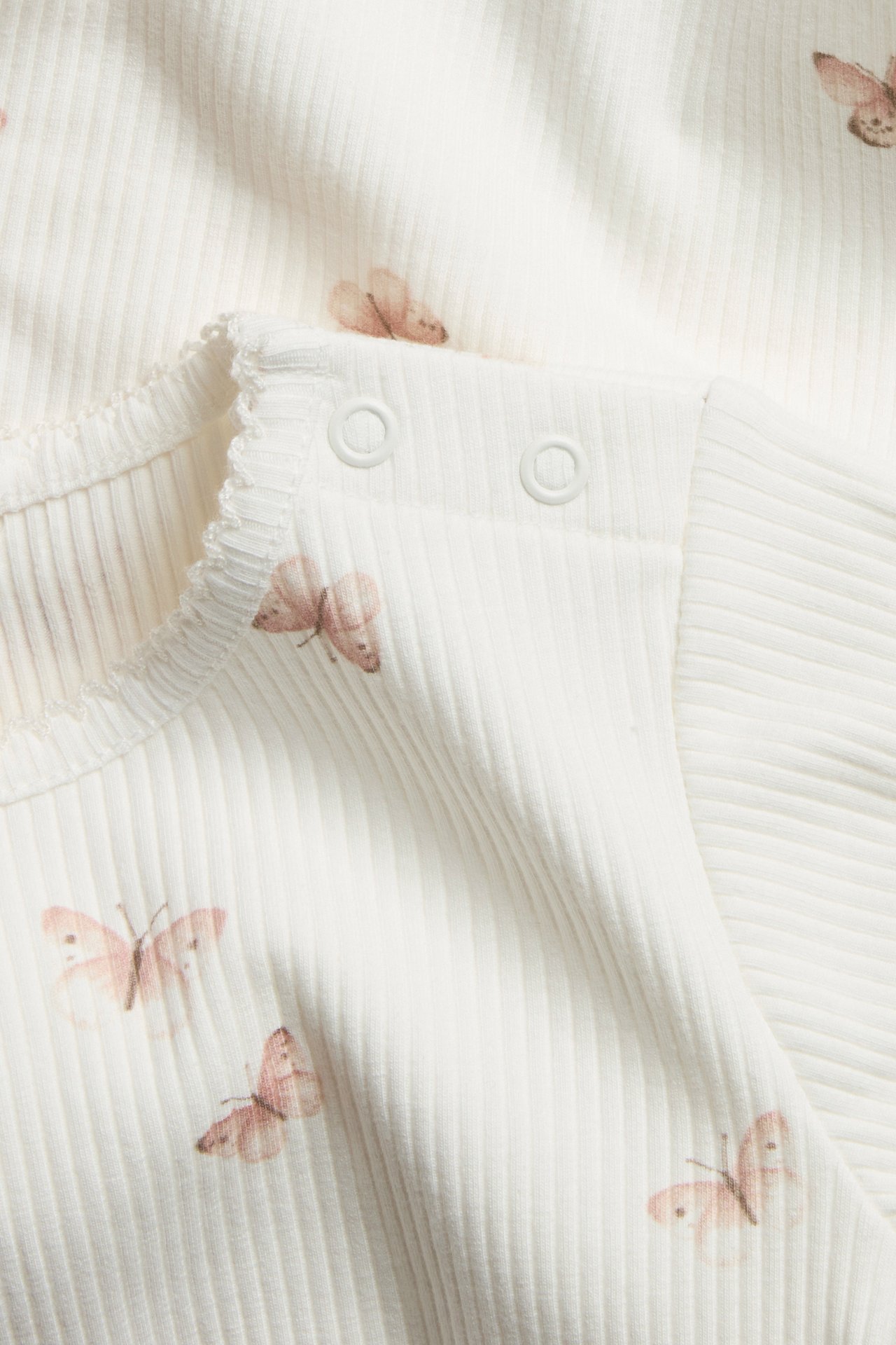 Nahaufnahme eines cremeweißen gerippten Baby-Bodys mit Schmetterlingsmuster und Schulter-Druckknöpfen.