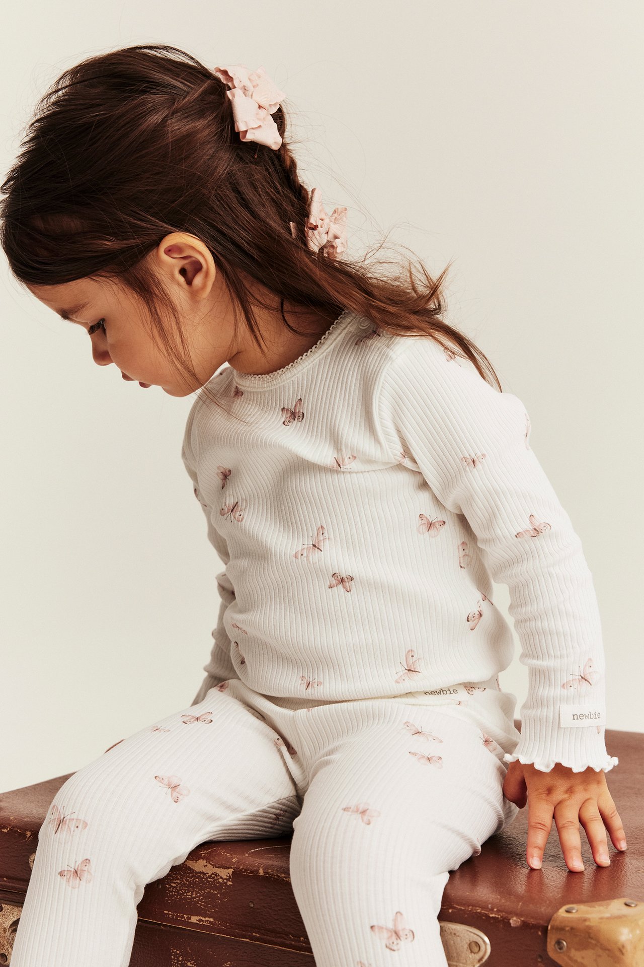 Seitenansicht: Kind trägt cremeweiße gerippte Schmetterlings-Leggings, ein passendes Oberteil und eine rosa Haarschleife.