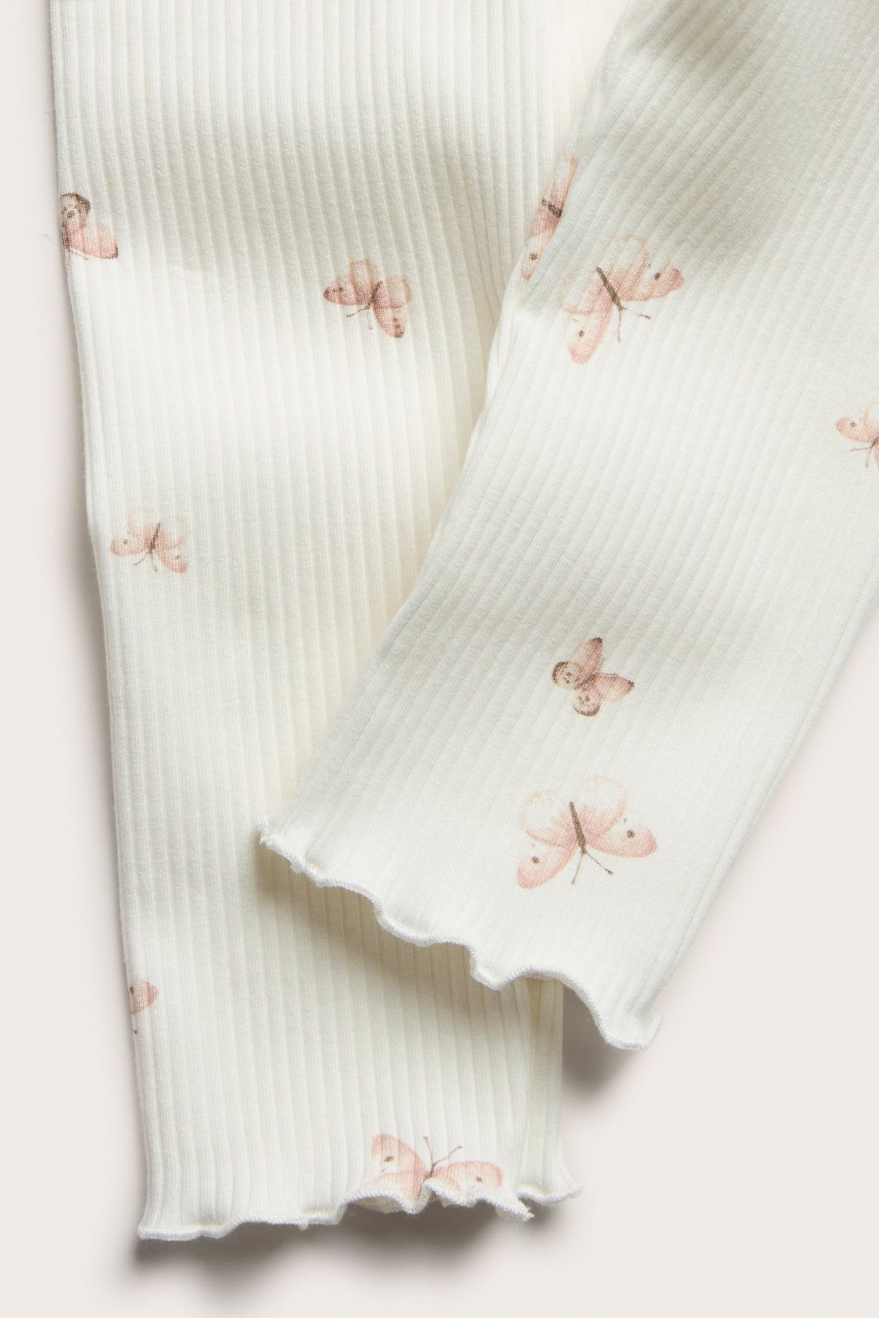 Nahaufnahme von cremefarbenen Ripp-Leggings mit hellrosa Schmetterlingsmuster und welligen Babylock-Säumen.