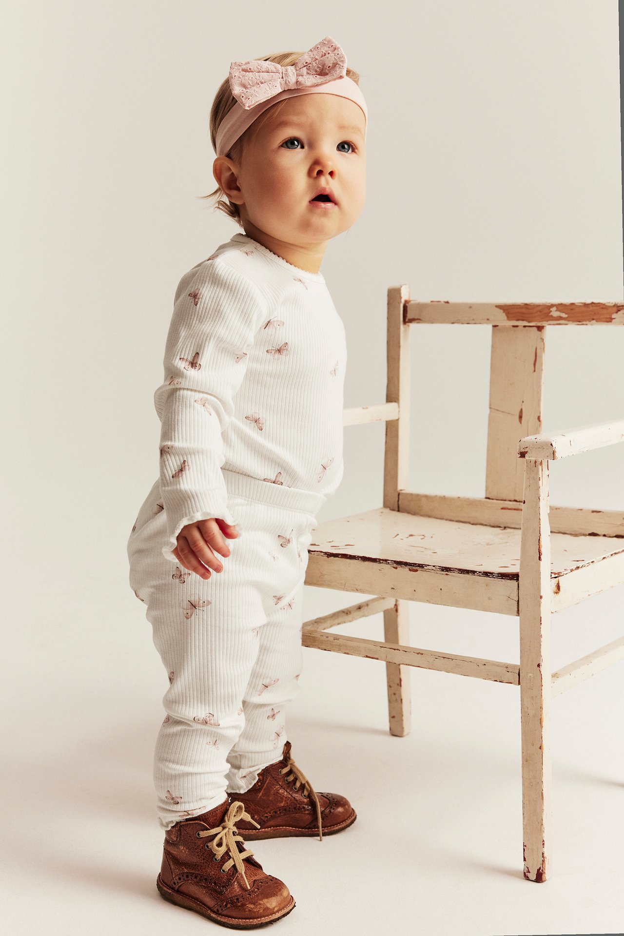 Side view of a baby in offwhite ribbed butterfly-patterned leggings, a matching top, light pink headband, and brown boots.