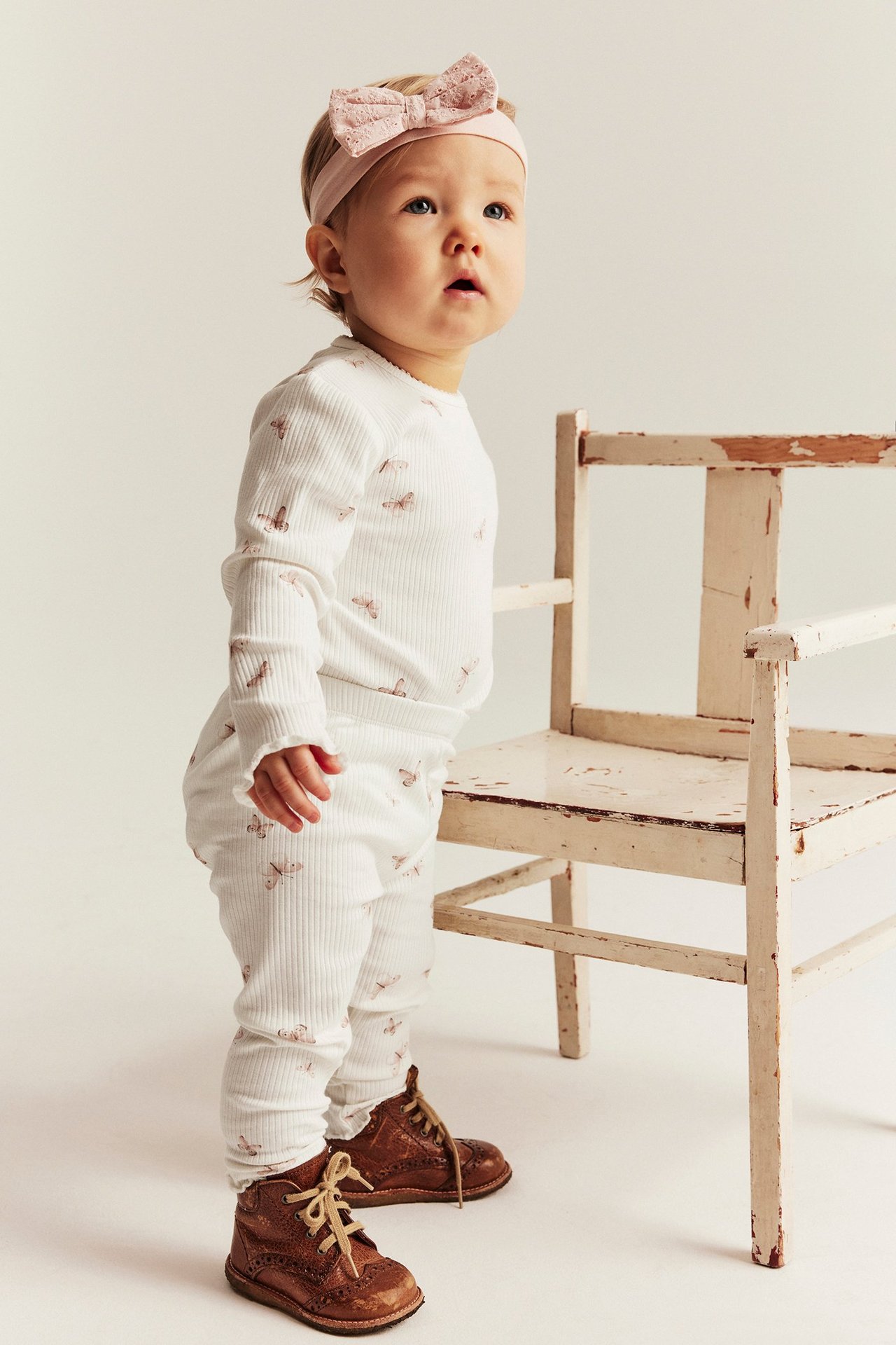 Front-side view of a baby wearing offwhite ribbed butterfly-patterned leggings, matching top, a pink bow headband, and brown shoes.