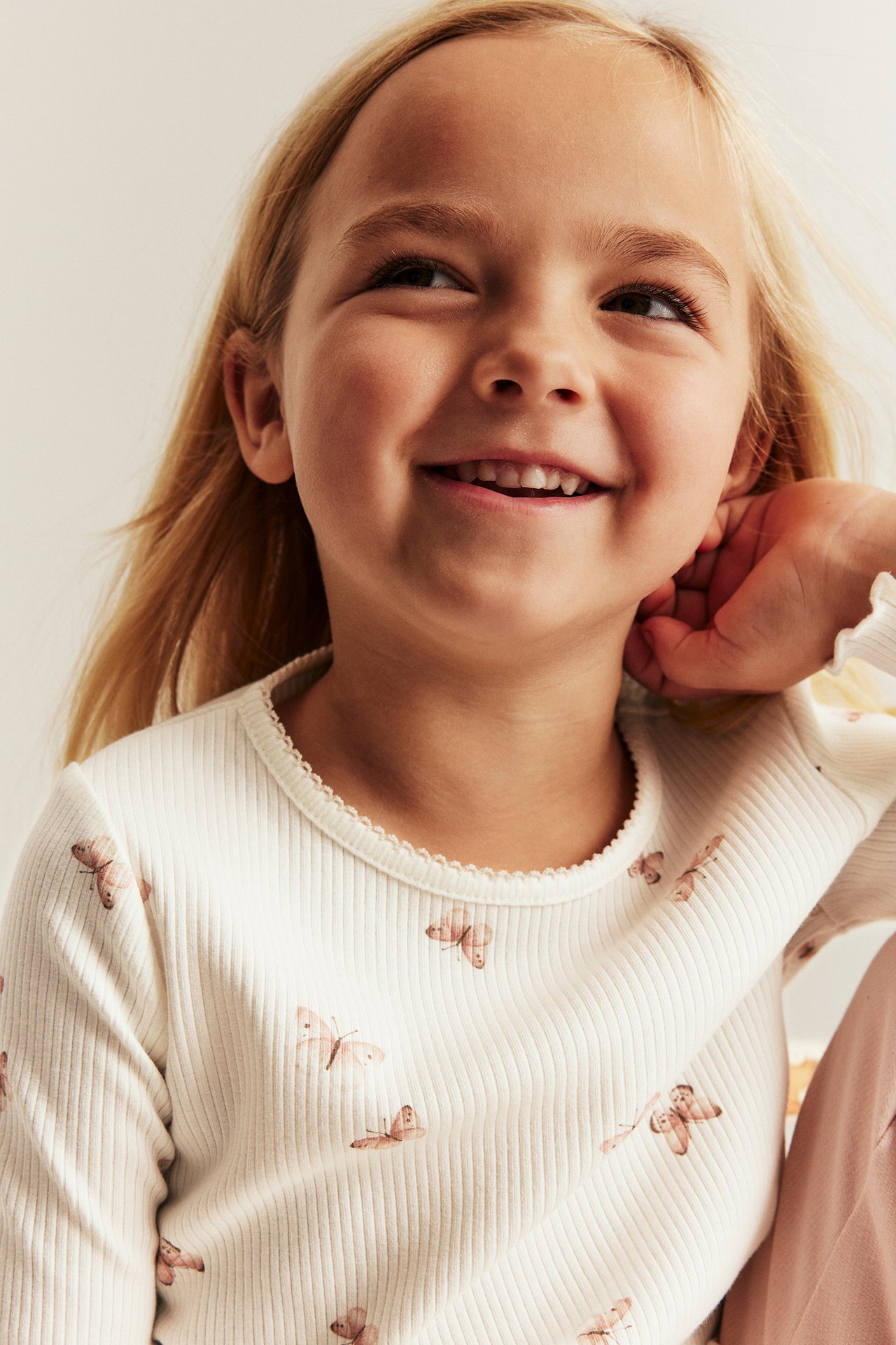 Nahaufnahme eines Kindes in einem cremefarbenen, gerippten Langarmshirt mit Schmetterlingsmuster und Picot-Saum.