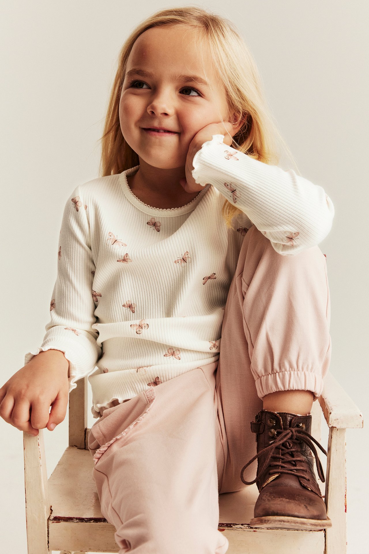 Frontansicht eines Kindes in einem cremeweißen gerippten Langarm-Top mit Schmetterlingsmuster, rosa Hose und braunen Stiefeln.