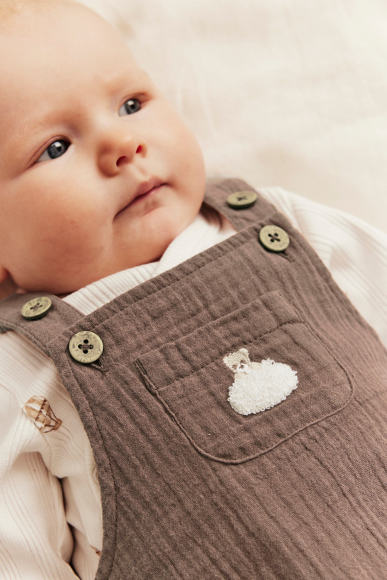 Close-up of baby in brown muslin overall with buttons and teddy bear embroidery over a white ribbed top.