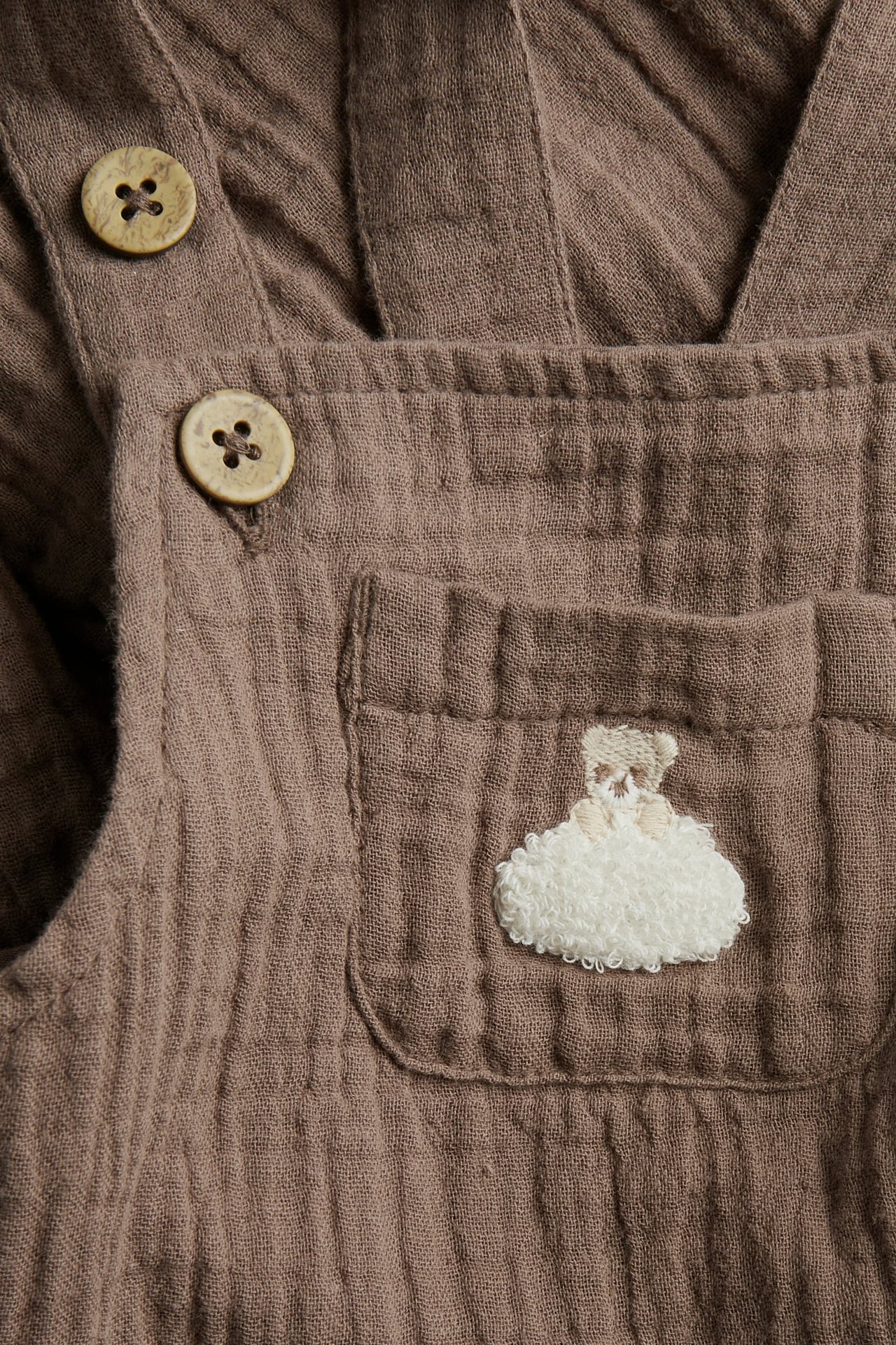 Close-up of brown muslin baby dungarees with wooden buttons and an embroidered teddy bear cloud.