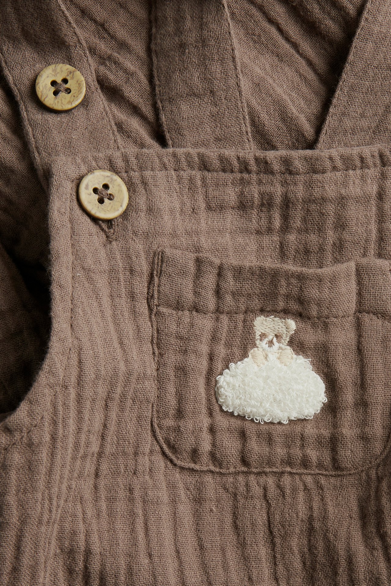 Close-up of brown muslin baby dungarees with wooden buttons and an embroidered teddy bear cloud.