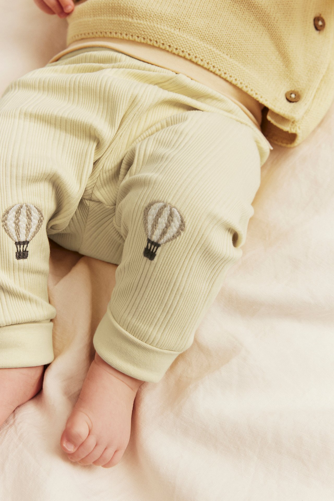 Nahaufnahme von Baby in gelben gerippten Leggings mit Heißluftballon-Applikationen und gelber Strickjacke.