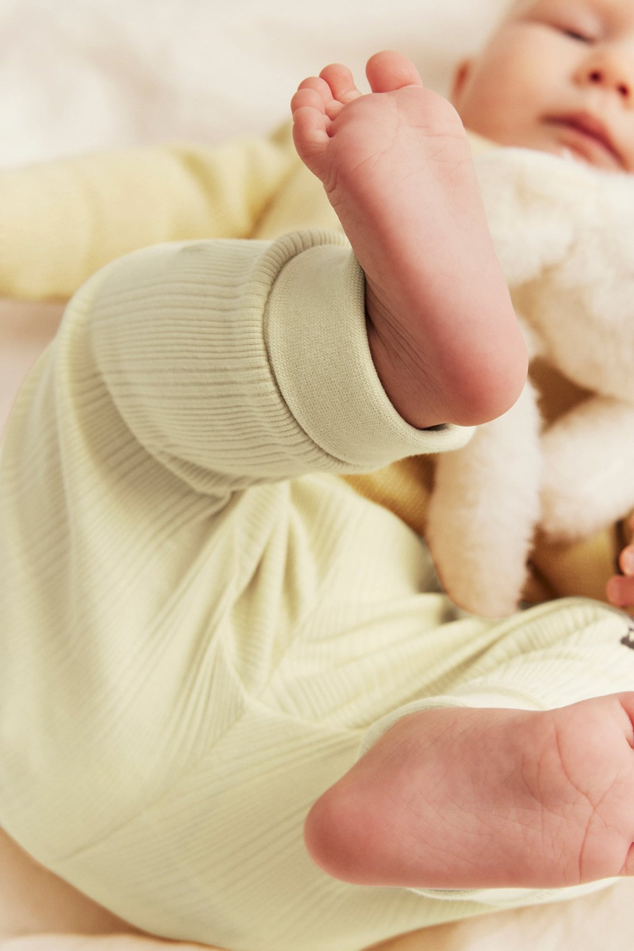 Nahaufnahme von Baby in hellgelben Ripp-Leggings mit Umschlägen, einem hellgelben Oberteil, das ein weißes Kuscheltier hält.