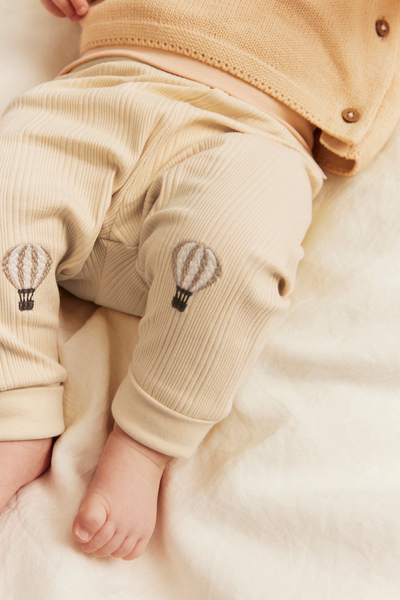 Nahaufnahme von Baby in gerippter beiger Hose mit Heißluftballon-Stickerei und beiger Strickjacke.