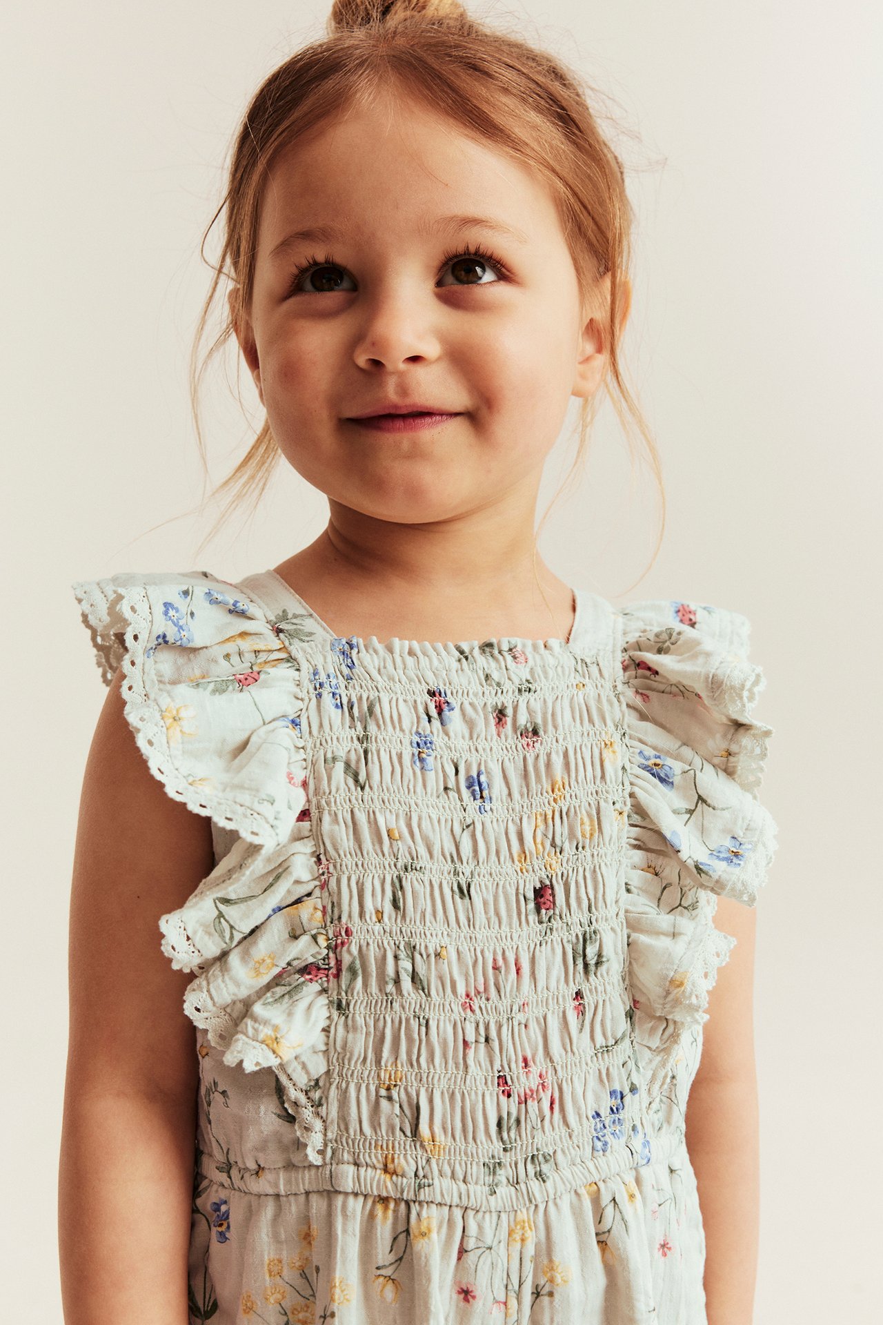 Close-up front view of a kid in a light green floral woven short jumpsuit with smocked elastic and ruffled straps.