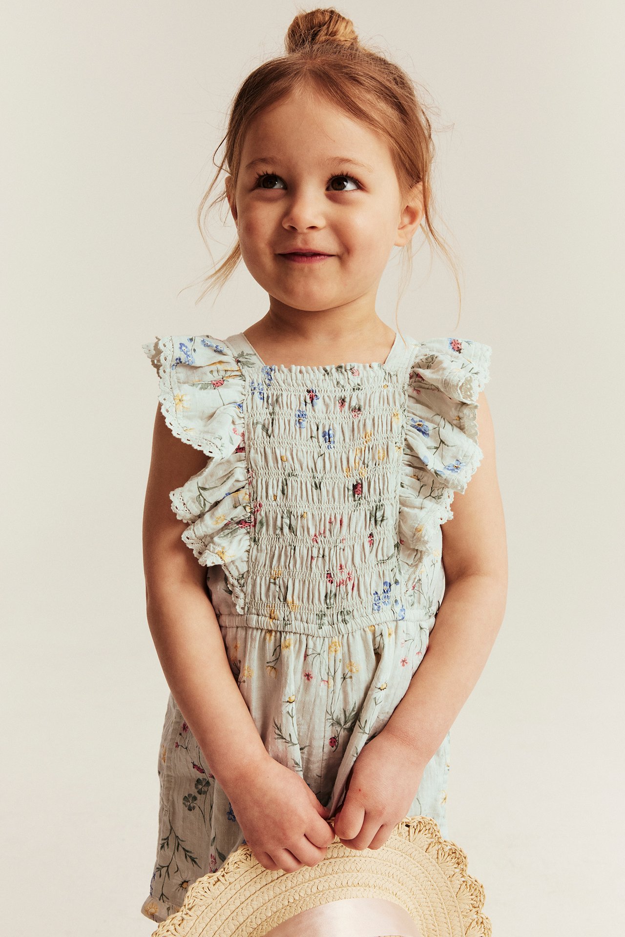 Front view of a kid wearing a light green floral smocked jumpsuit with ruffles, holding a straw hat.