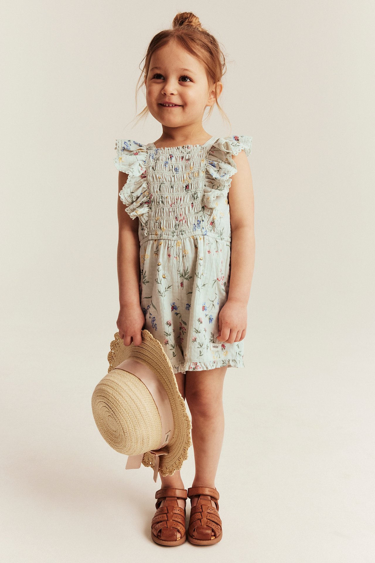 Front view: Kid in light green floral short jumpsuit with smocking, ruffles. Holds straw hat, wears brown sandals.