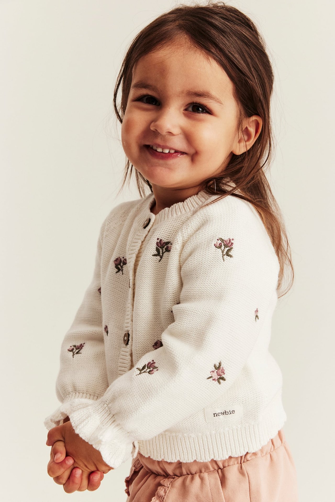 Lächelndes Kind in cremeweißer Strickjacke mit Blumenstickerei und rosa Rüschenhose, Frontansicht.