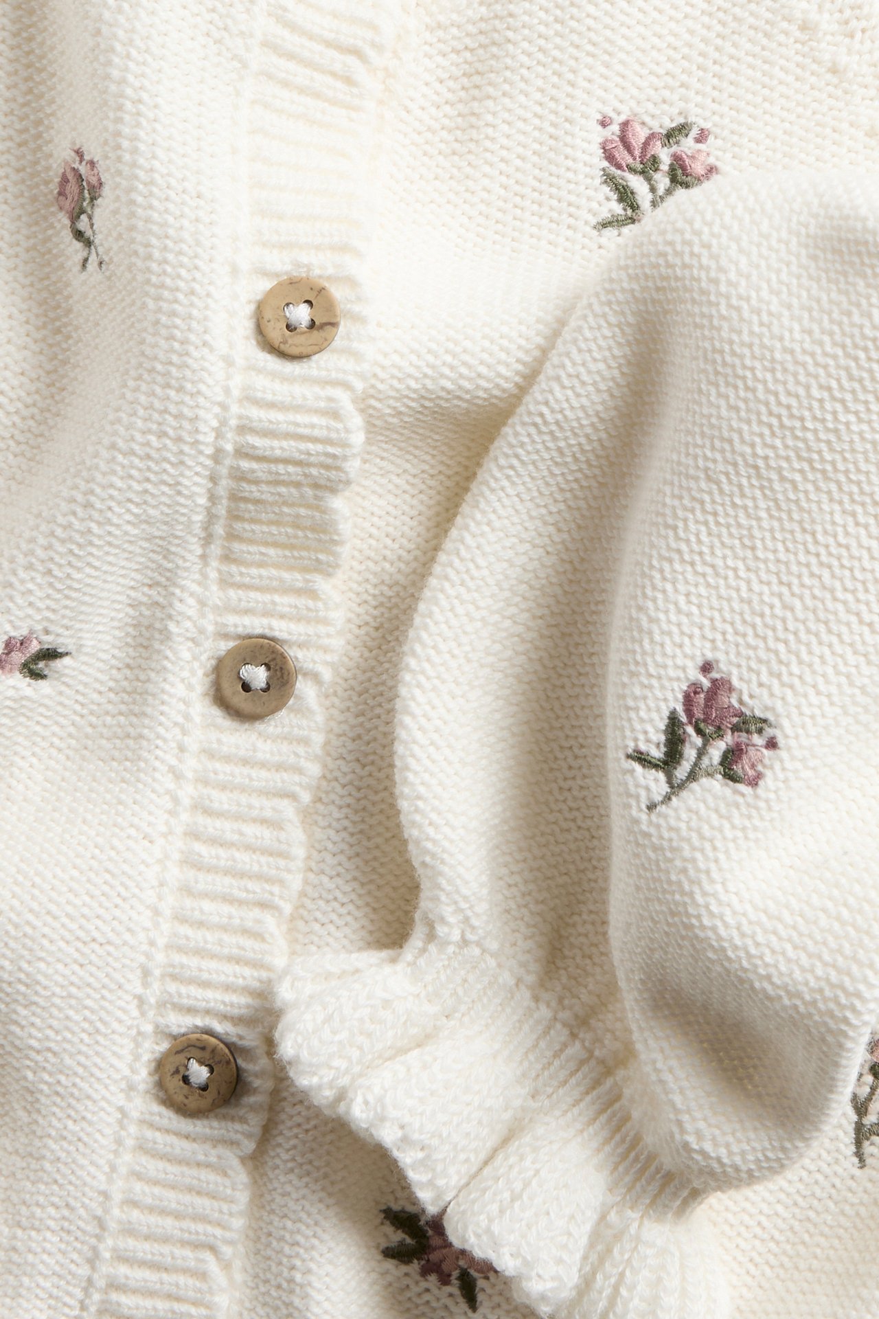 Nahaufnahme cremeweißer Baumwoll-Strickjacke mit gestickten rosa Blumen, Holzknöpfen und Rüschen.