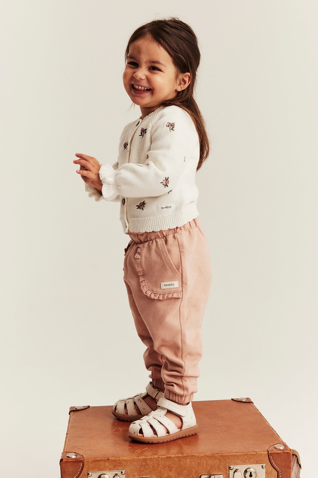 Lächelndes Kind in einer cremeweißen bestickten Blumenstrickjacke, rosa Rüschenhose und weißen Sandalen. Seitenansicht.