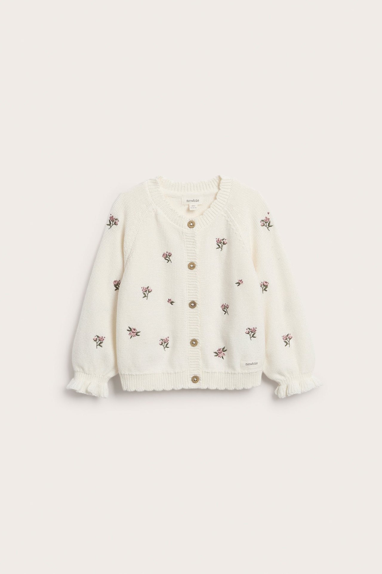 Vorderansicht einer cremeweißen gestrickten Kinder-Baumwollstrickjacke mit rosa Stickblumen, Knopfleiste, Wellensaum und Rüschenbündchen.