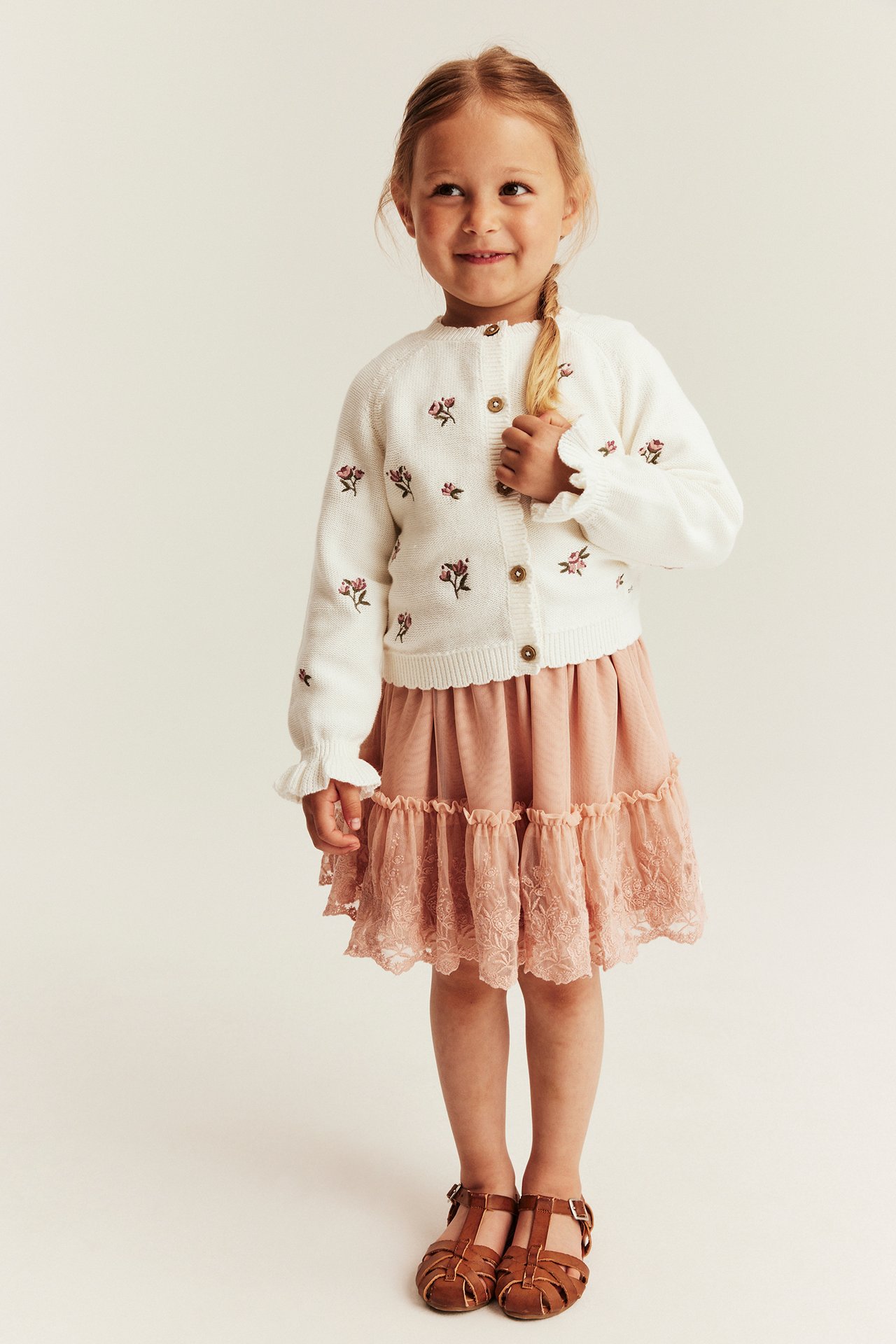 Front view of a kid wearing an offwhite knitted cotton cardigan with embroidered flowers, a pink tiered skirt, and brown sandals.