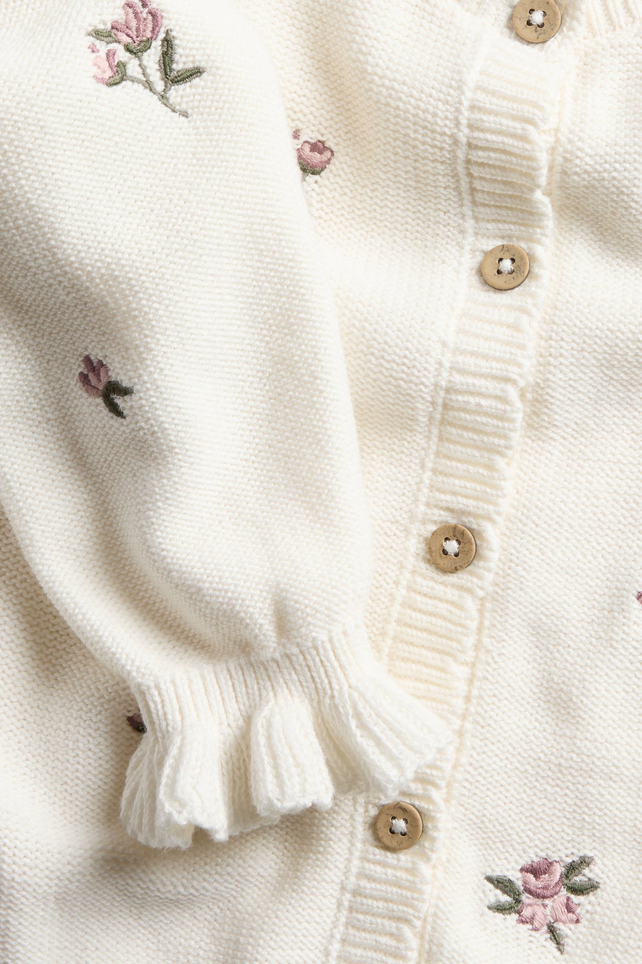 Close-up of an off-white cotton knitted cardigan with pink embroidered flowers, ruffled cuffs, and wooden buttons.