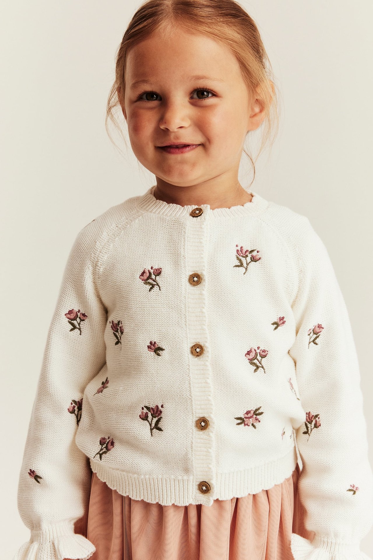 Vorderansicht: Kind in cremefarbenem Baumwollstrick-Cardigan mit rosa Stickblumen, Holzknöpfen und rosa Rock.