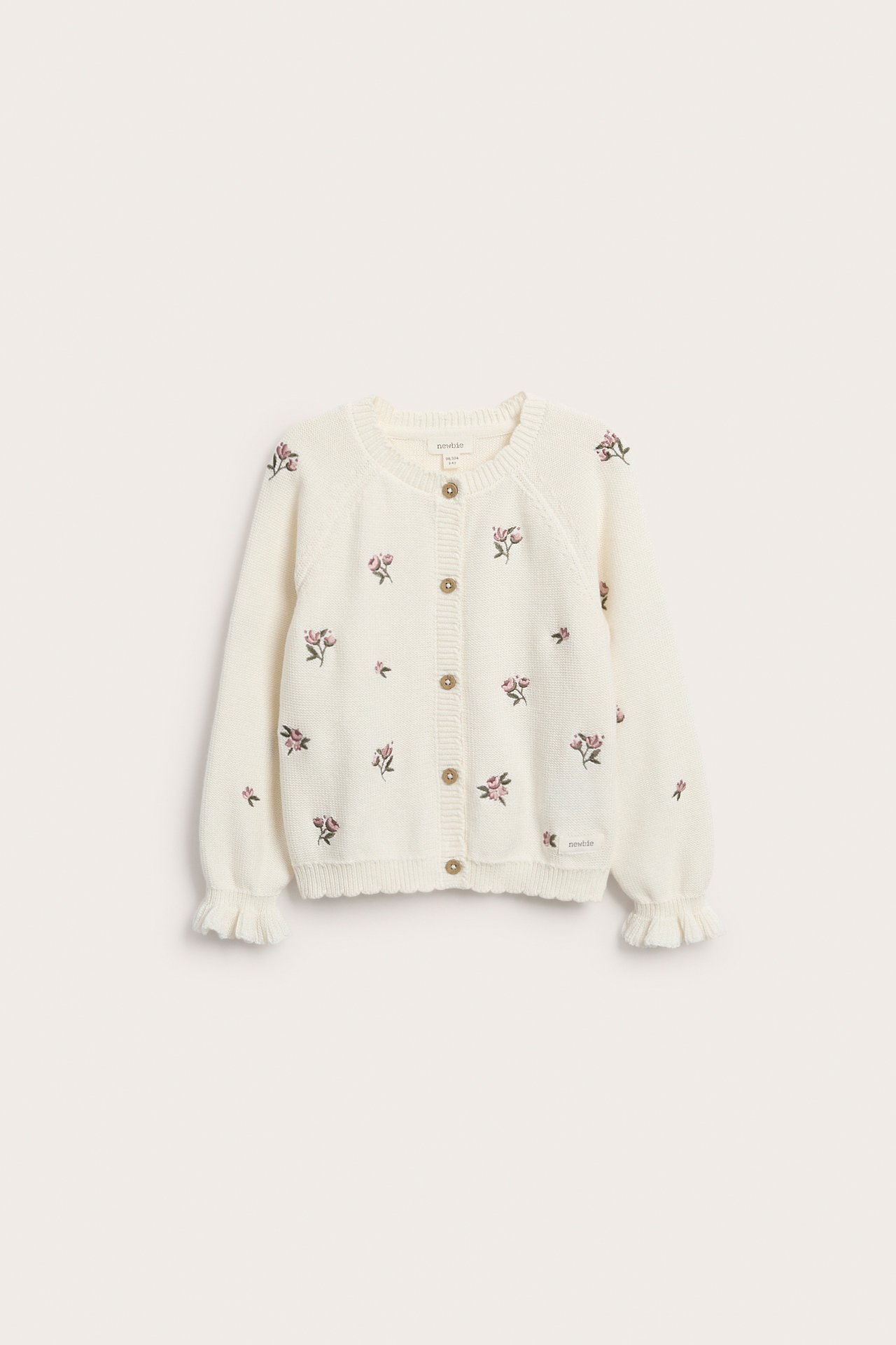 Front view: Off-white knitted cotton cardigan with embroidered pink flowers, wooden buttons, and ribbed scalloped cuffs.