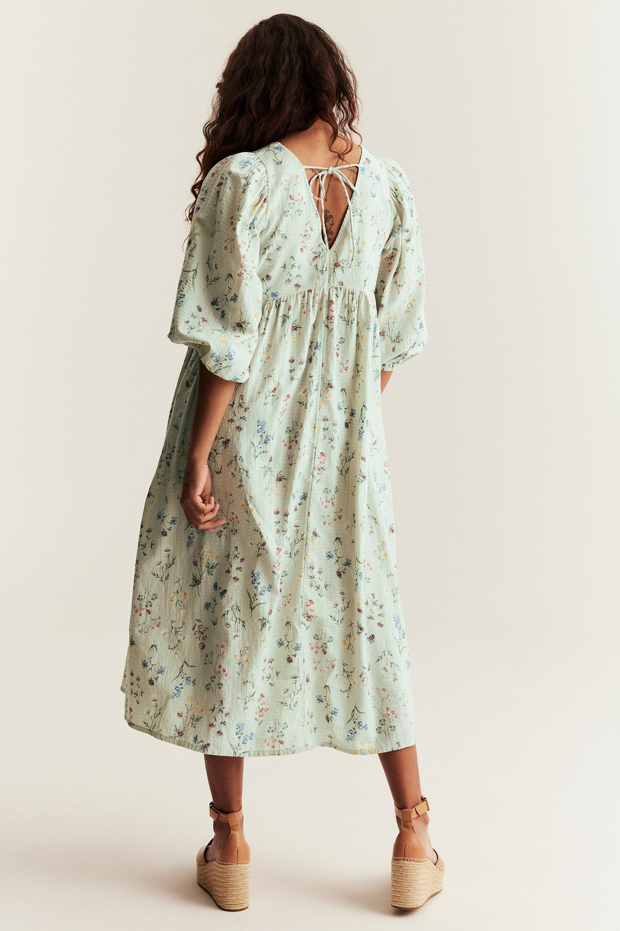 Back view of woman in light green floral cotton midi dress, puff sleeves, tie-back, and wedge sandals.