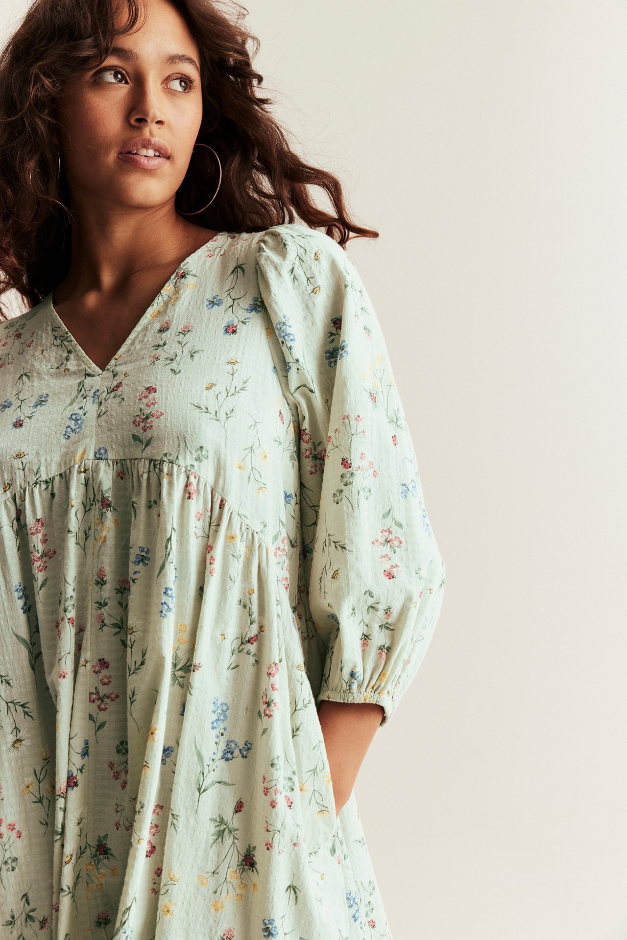 A woman wears a light green floral cotton V-neck dress with puff sleeves and hoop earrings.