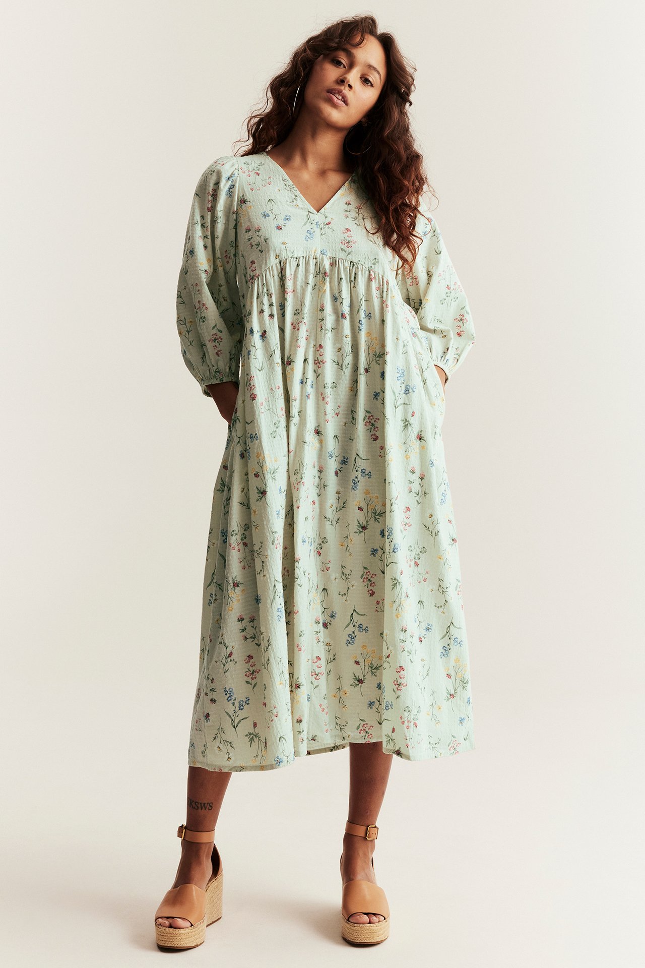 Front view: Woman wearing a light green floral cotton V-neck midi dress, puff sleeves, and wedge sandals.