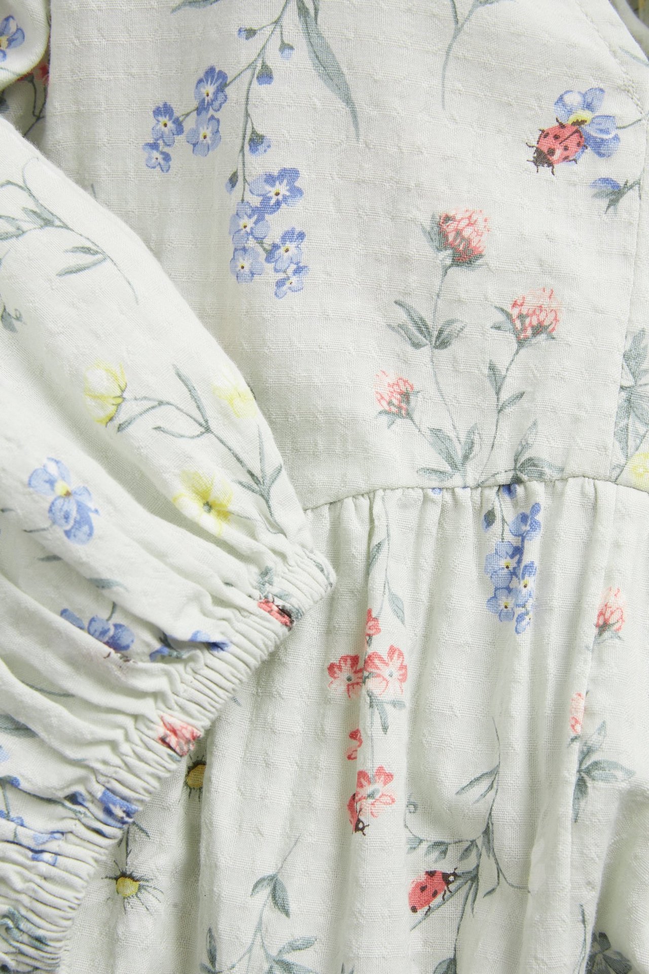 Close-up: Light green cotton dress with floral pattern featuring blue, pink flowers and red ladybugs.