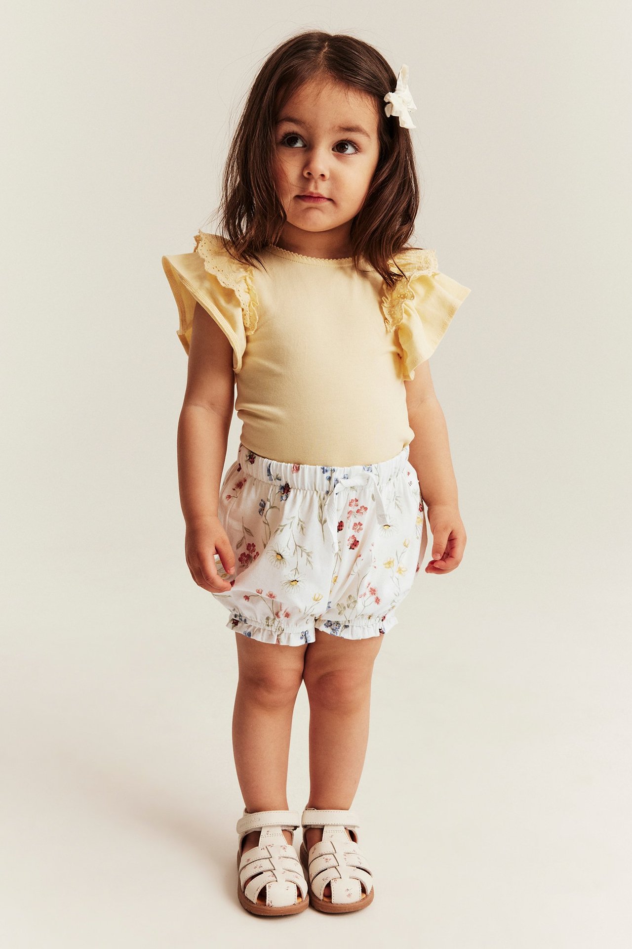 Vorderansicht eines Kindes in weißen geblümten Jersey-Shorts mit Rüschen, einem gelben Oberteil und hellen Sandalen.