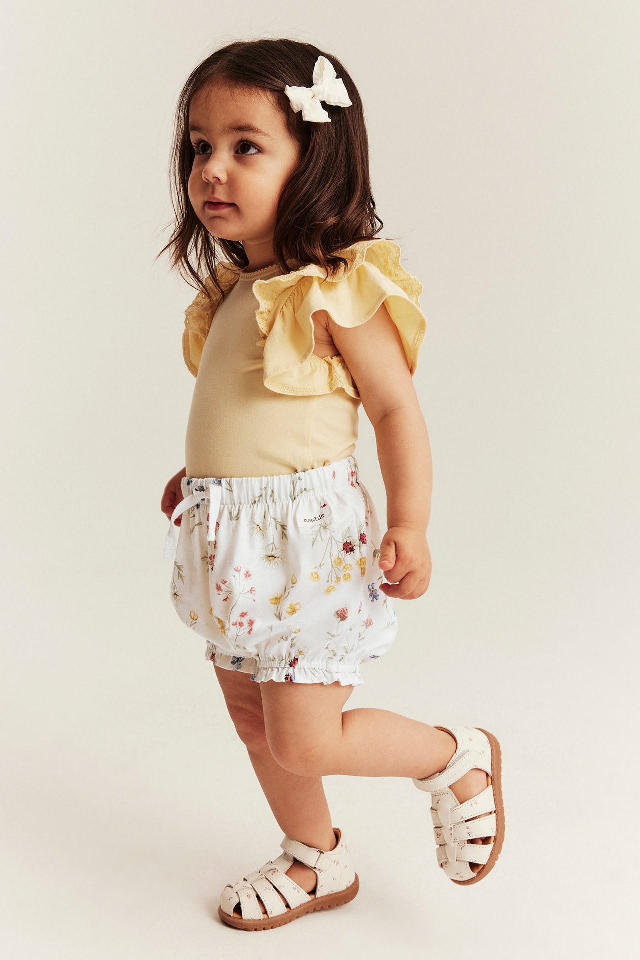 Seitenansicht eines Babys in ecrufarbenen geblümten Puff-Shorts, gelbem Rüschenoberteil und weißen Sandalen.