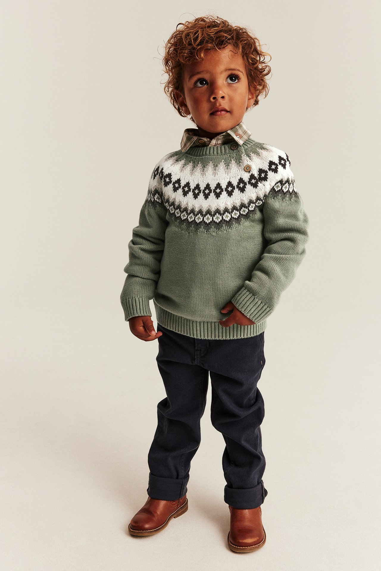 Front view: kid in green Fair Isle sweater, plaid shirt, dark blue pants, brown boots.