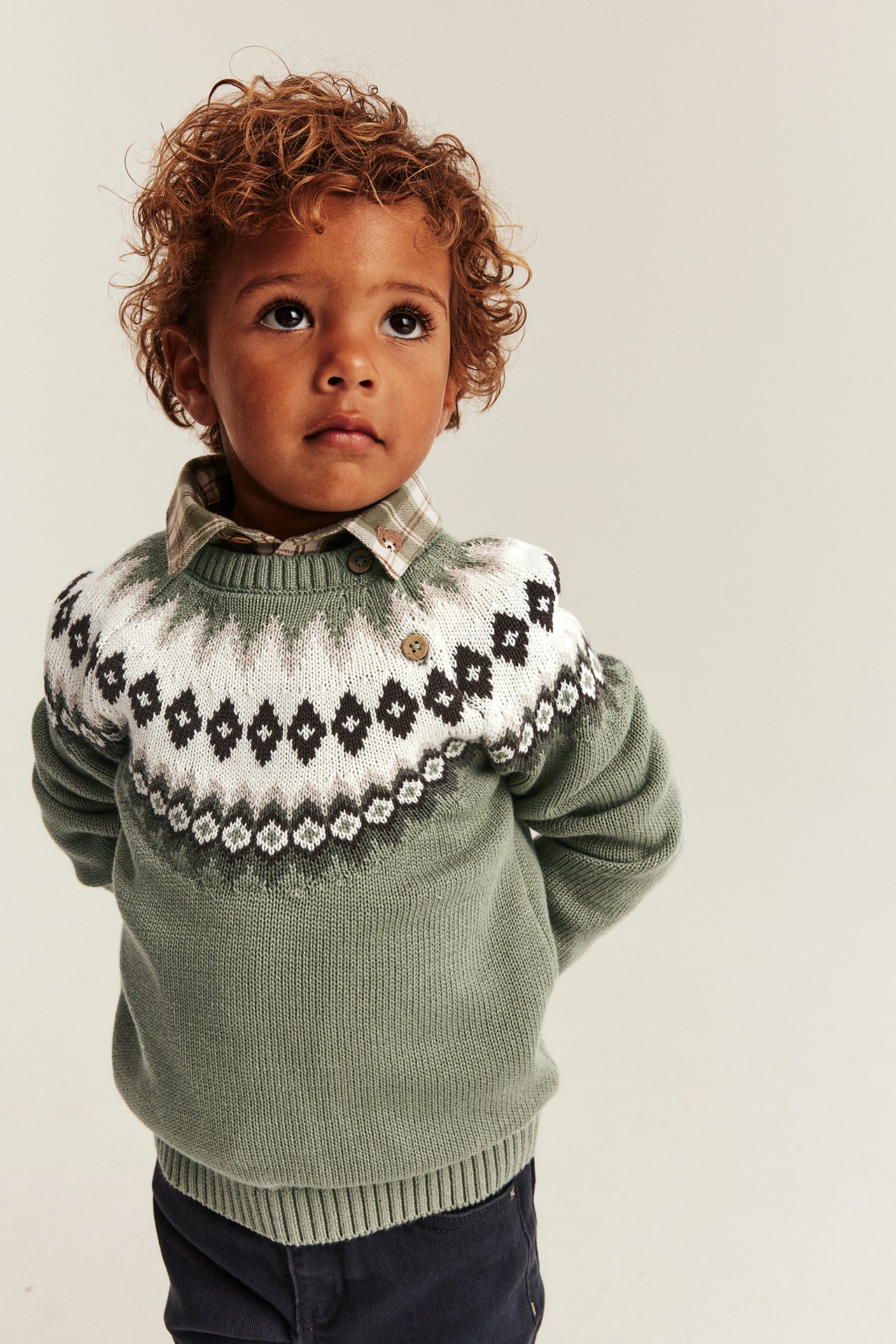 Frontansicht: Kind in grünem Fair-Isle-Strickpullover über kariertem Hemd und dunkler Hose.