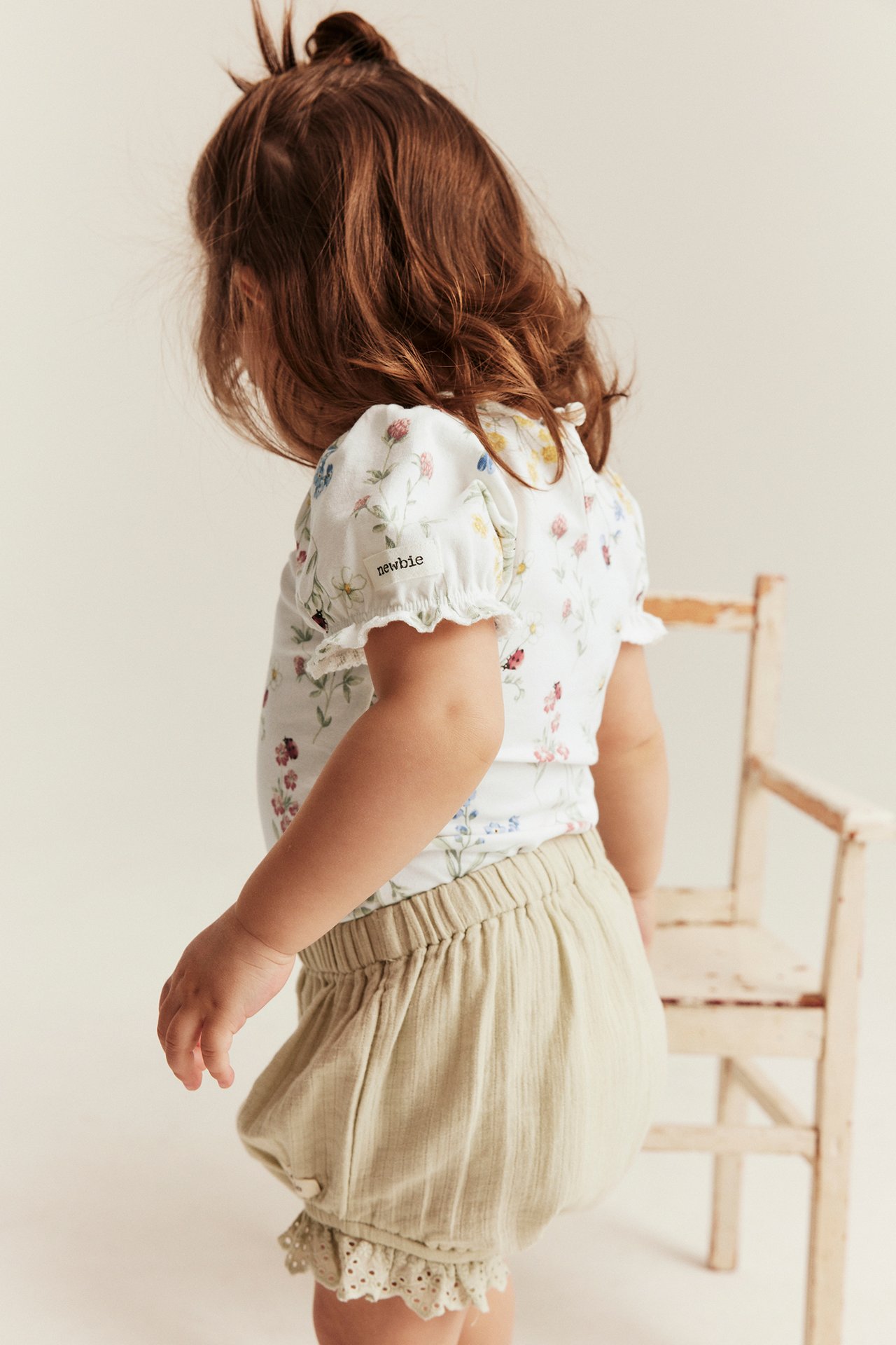 Back view of a baby in an off-white floral puff-sleeve bodysuit and light beige ruffled shorts.