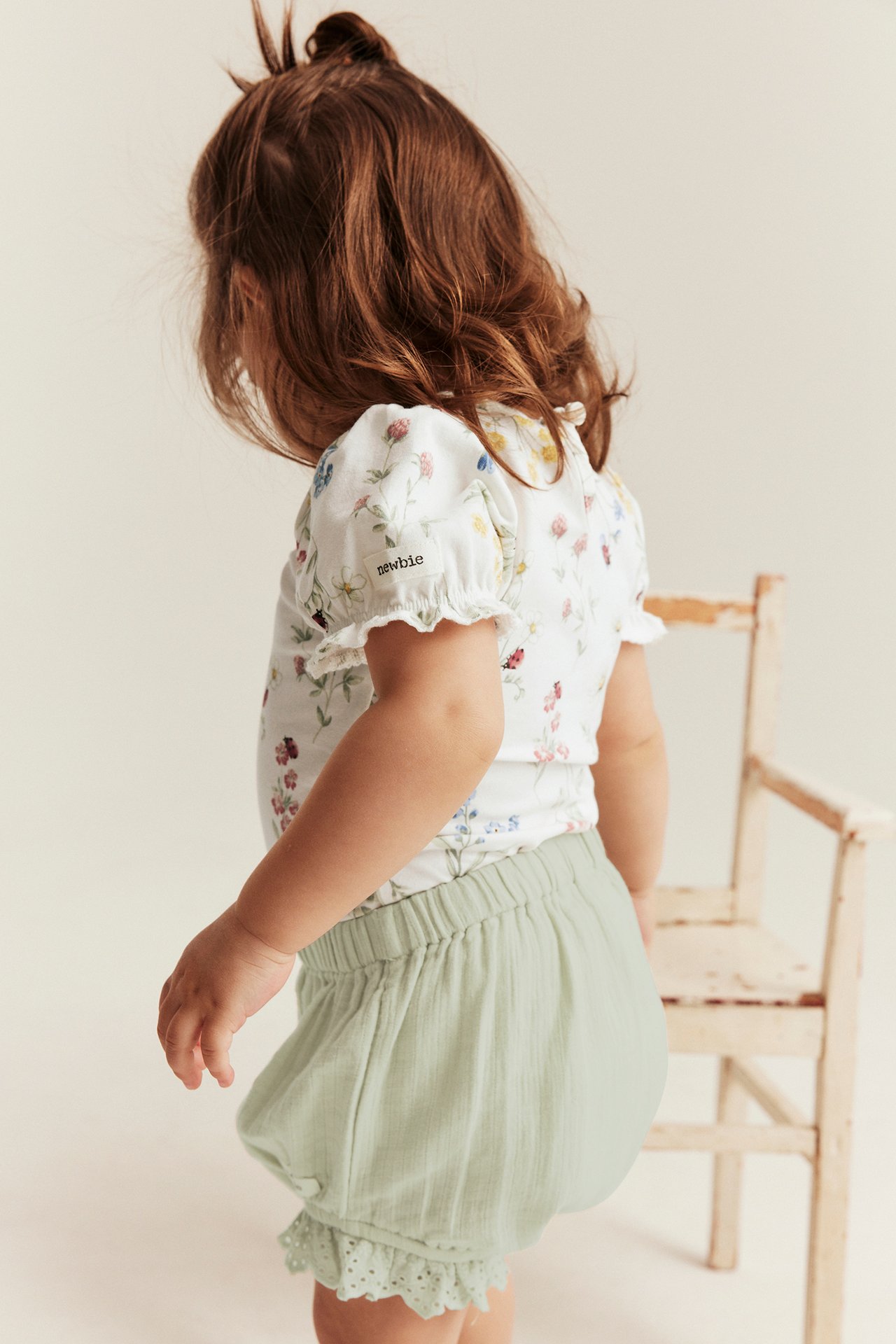 Back view of baby in off-white floral short-sleeved bodysuit with puff sleeves and light green ruffled shorts.