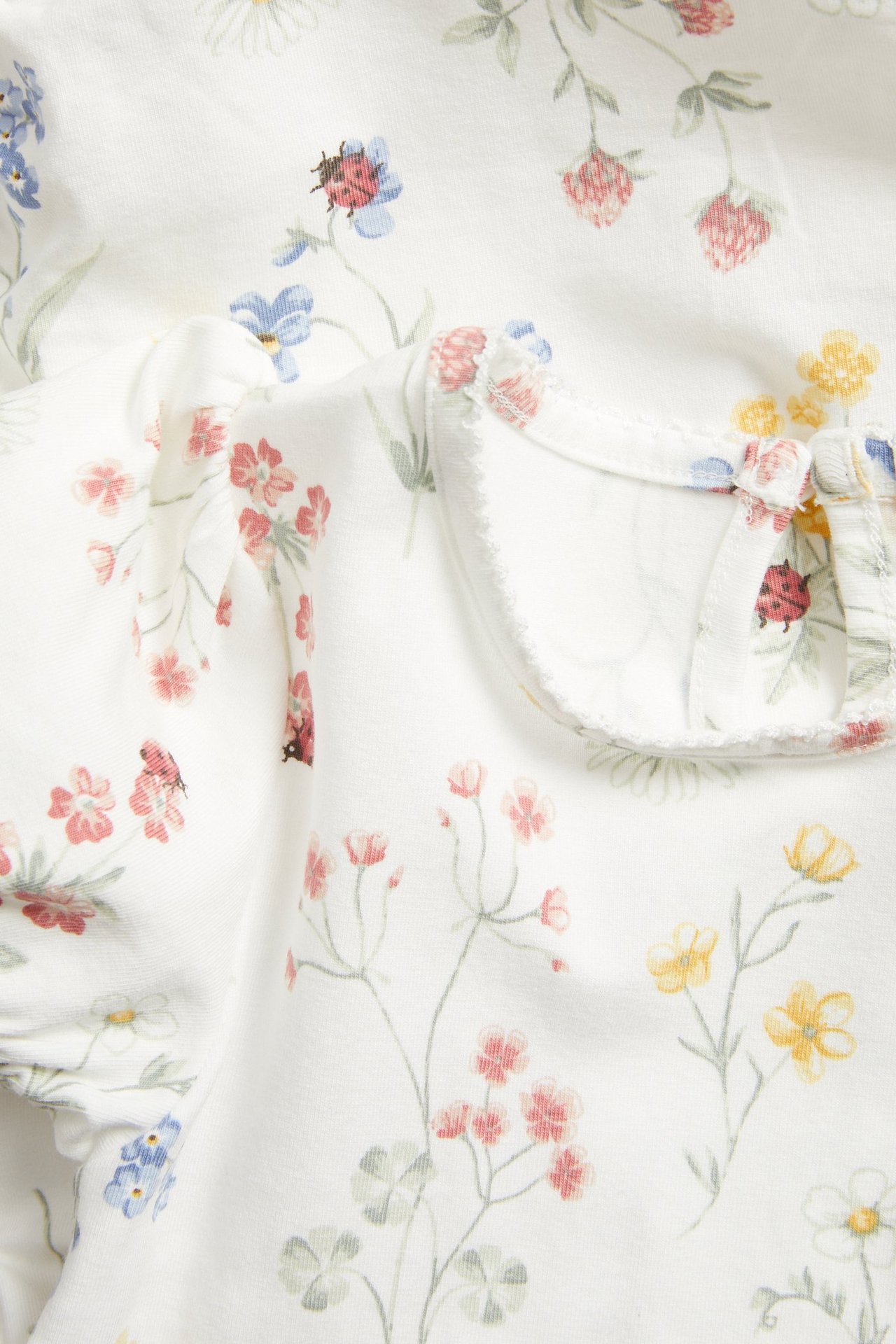Nahaufnahme eines cremeweißen Baby-Bodys mit Blumen- und Marienkäferdruck, kurzen Puffärmeln und Picot-Saum.