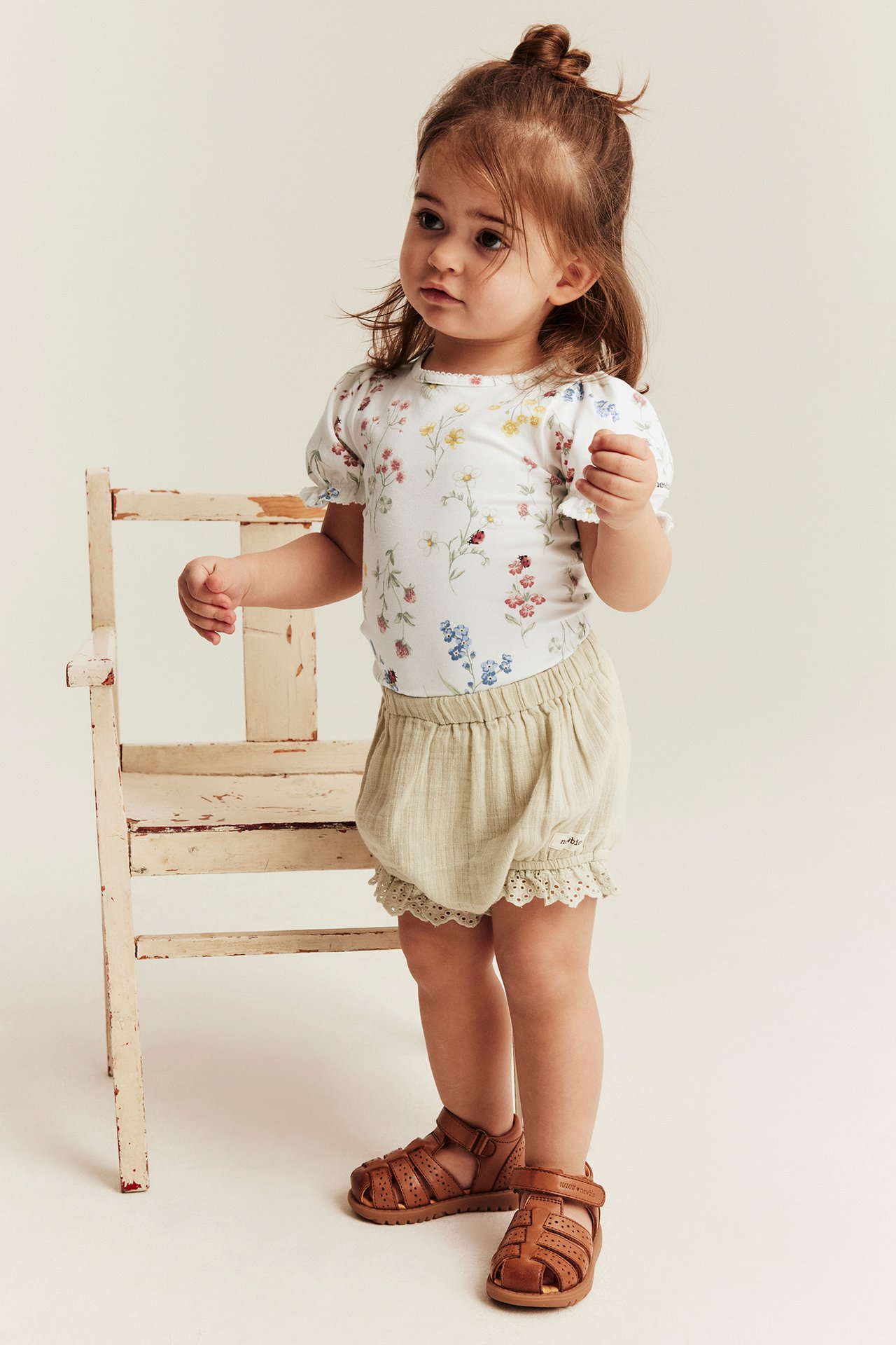 Front view of baby in an offwhite floral short-sleeved bodysuit, light beige shorts with lace, and brown sandals.