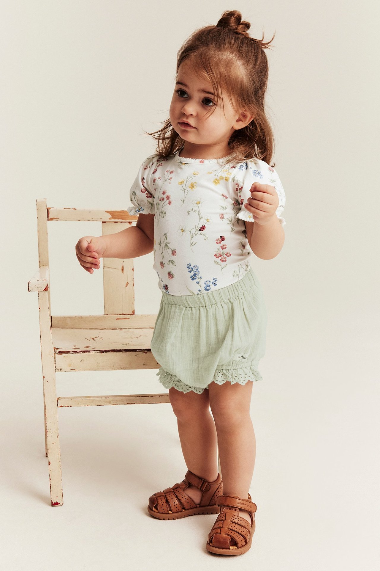 Front view: Baby wears off-white floral jersey bodysuit, light green crinkle shorts, brown sandals.