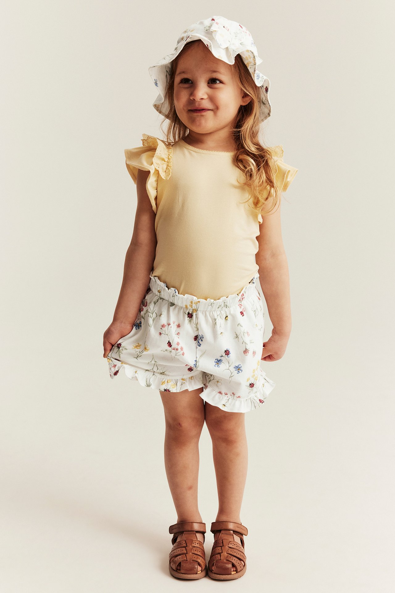 Front view: Kid wears offwhite floral jersey shorts with ruffles, yellow top, floral hat, and brown sandals.